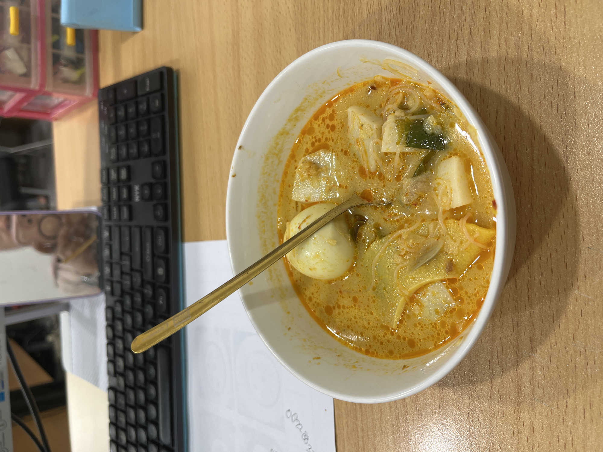 A bowl of laksa is placed on a wooden desk next to a keyboard. The laksa contains noodles, tofu, and a hard-boiled egg, with a gold spoon resting in the bowl. In the background, a computer monitor and a stack of pink plastic drawers are visible. The overall scene suggests a workspace environment, possibly in an office, with the food indicating a meal break. The image does not provide any specific visual cues that would allow for the identification of the city of Pekanbaru, Indonesia.