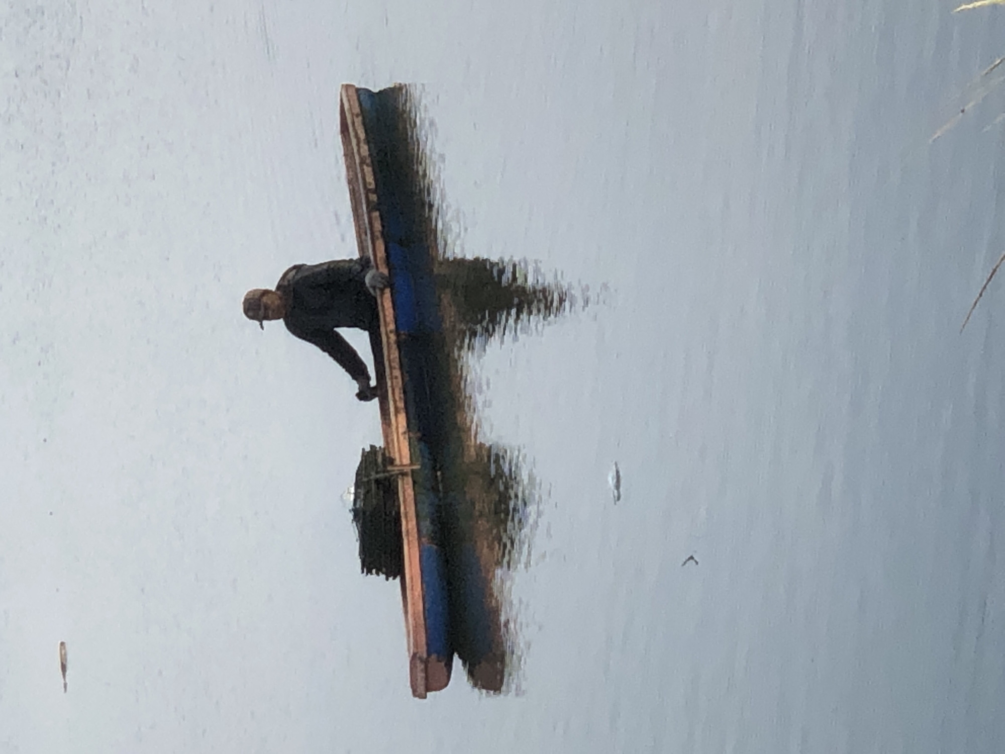 A person wearing a cap and dark clothing is sitting in a long, narrow boat on a body of water. The boat has a blue hull with a wooden interior and is carrying a stack of green, square objects. The person appears to be rowing. The water is calm and reflects the boat and the person. There are a few small objects or debris floating on the surface of the water. In the distance, a small, light-colored object is visible on the water, possibly a buoy or a bird. The overall scene appears to be a quiet, natural environment. The image does not provide specific landmarks or details to definitively place it in Lhokseumawe, Indonesia, but such scenes are common in coastal areas and rivers of Indonesia.