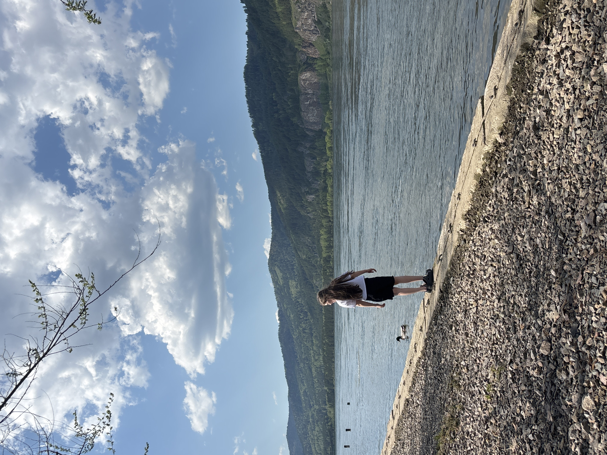 A young woman stands on a concrete embankment alongside a river in Овсянка, Russia. She wears a white shirt and black skirt. The river is wide and calm, with a mountain visible in the background. The sky is blue with scattered clouds. To the right of the embankment is an area covered in small stones. A duck floats near the edge of the embankment.