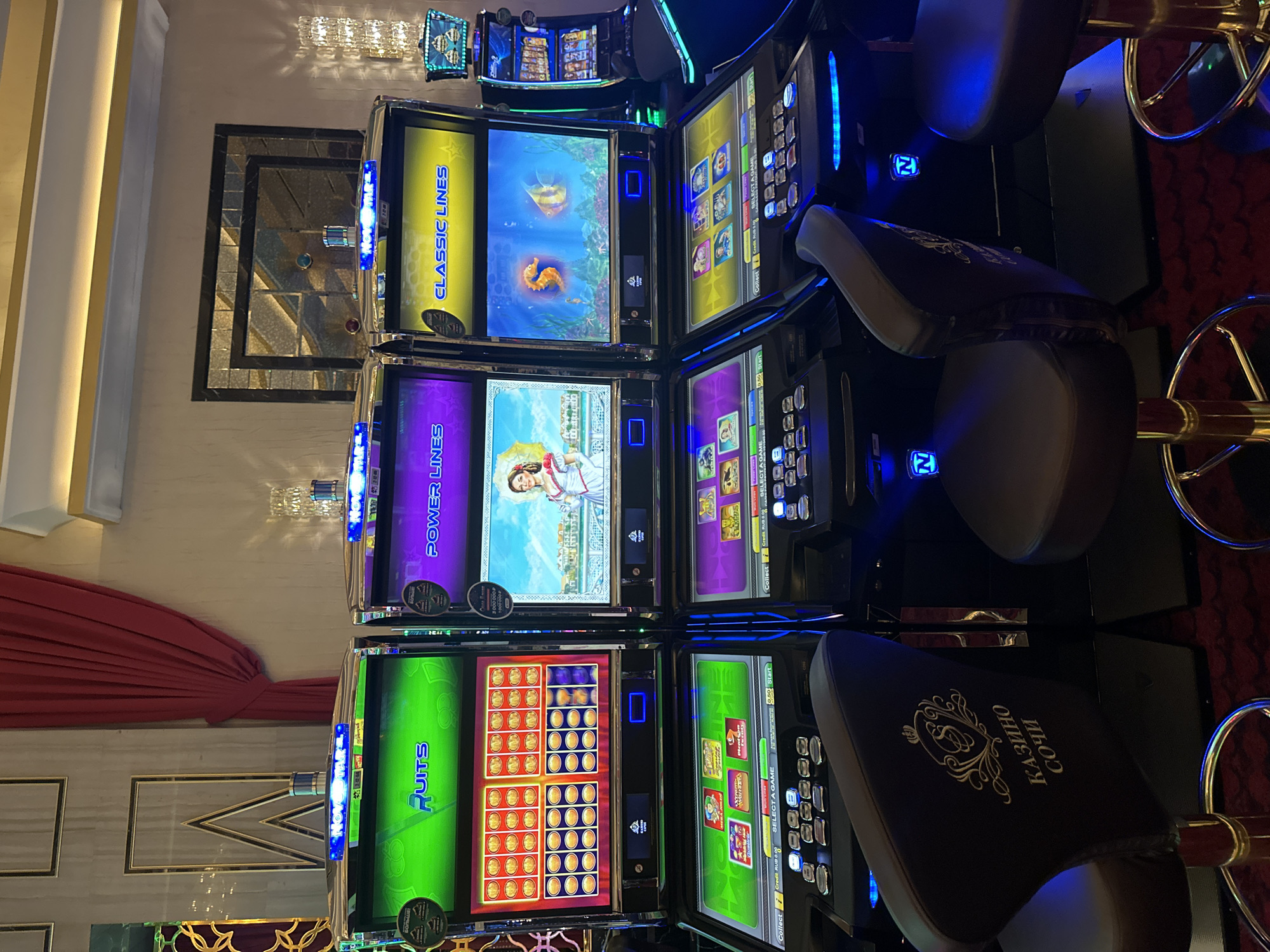 The image shows a row of slot machines in a casino. The machines are brightly lit with flashing lights and colorful displays. The slot machines are labeled "Fruits", "Power Lines", and "Classic Lines". There are also chairs in front of the machines, with one chair in the foreground bearing the logo "Казино Сочи" (Casino Sochi). The environment appears to be a modern casino interior with ornate decorations and dim lighting. The casino is located in Estosadok, Russia, which is a resort town in the Krasnodar Krai region.