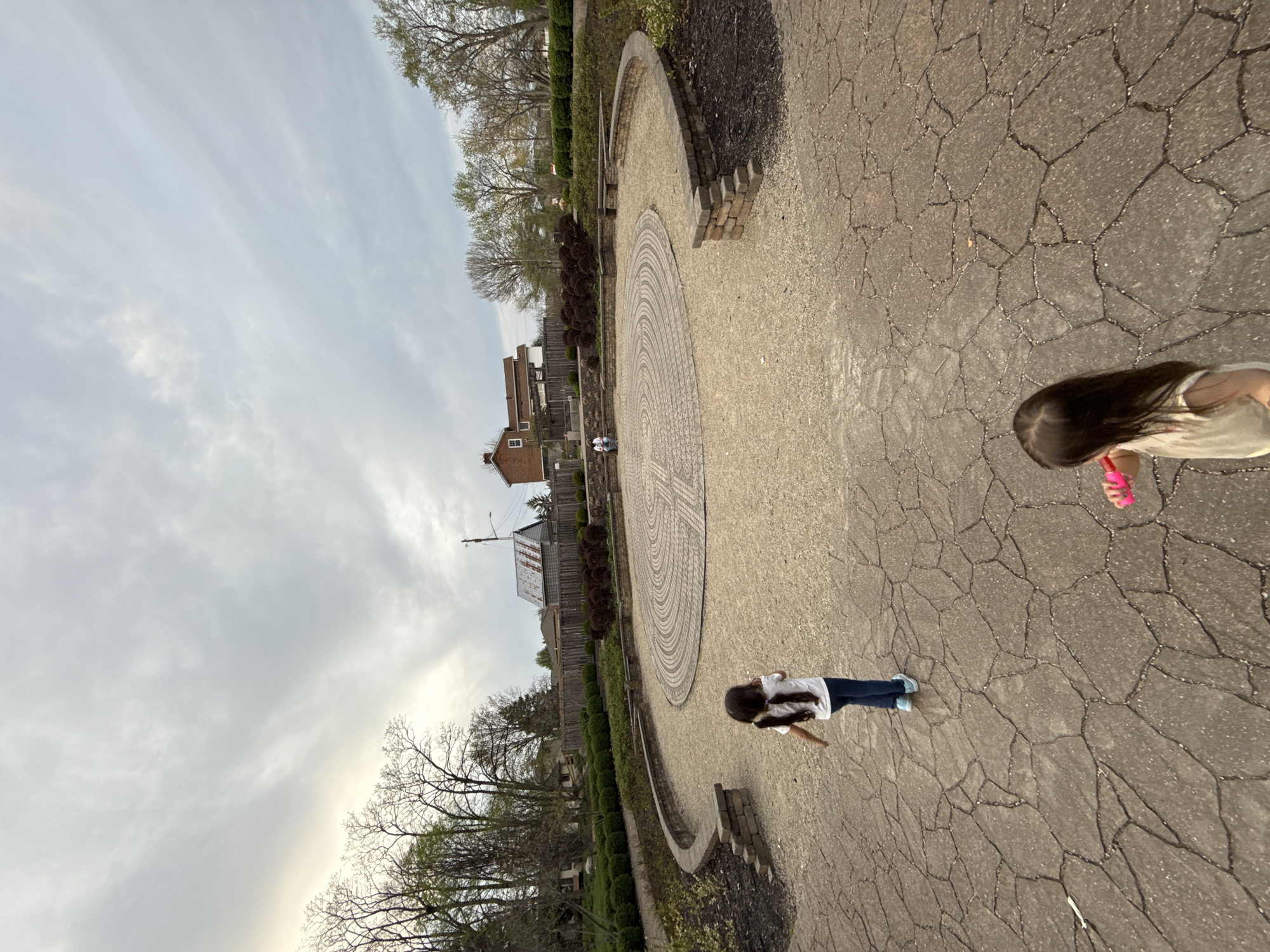 The image depicts an outdoor scene in Grove City, Ohio, United States of America. The setting appears to be a park or garden with a circular pattern inlaid in the pavement. Two children are visible on the cracked pavement. One is wearing a white shirt and blue jeans, facing the circular pattern. Another child, wearing a light-colored shirt and holding a pink object, is partially visible in the upper right corner. The environment includes buildings, trees, hedges, and cloudy skies.