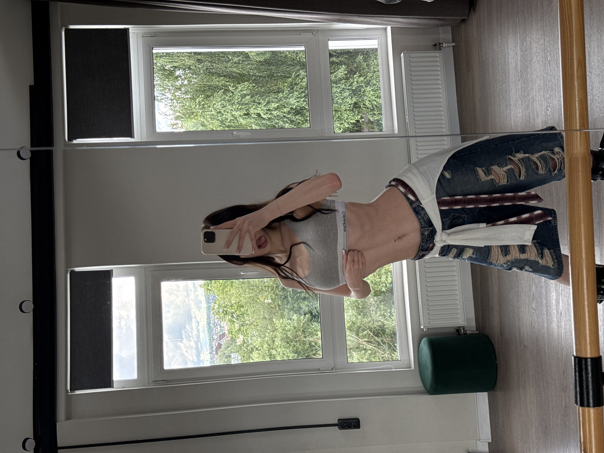 A young woman is reflected in a large mirror, posing with her tongue out. She is wearing a grey sports bra and distressed denim shorts, with a white shirt tied around her waist. The room features large windows with a view of trees and buildings, and a dark green ottoman is visible. A wooden barre is in the foreground. The scene is located in a room in Pokrovskoe, Russia.