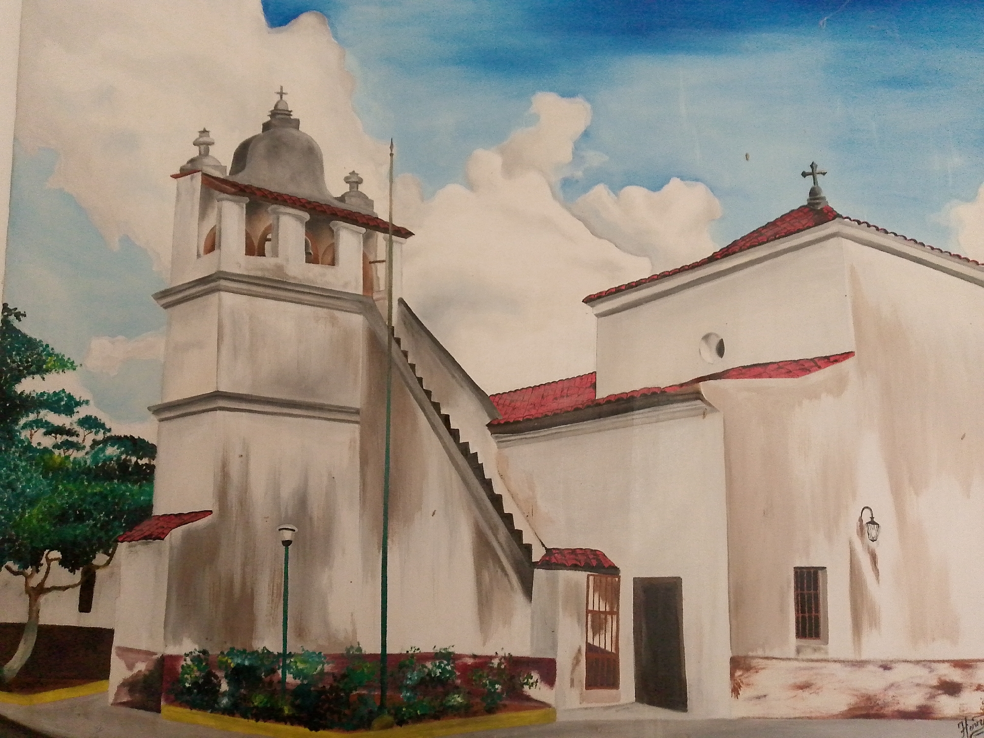 This is a painting of a Spanish colonial-style building, likely a church or mission, in Altagracia, Venezuela. The building is whitewashed with a red-tiled roof. A prominent bell tower with arched openings is to the left, topped by a dome. An exterior staircase leads up the side of the building. There is greenery, including trees and bushes, in the foreground. The sky is blue with white clouds. There are no people depicted in the painting. The overall impression is one of historical architecture and a bright, sunny day.