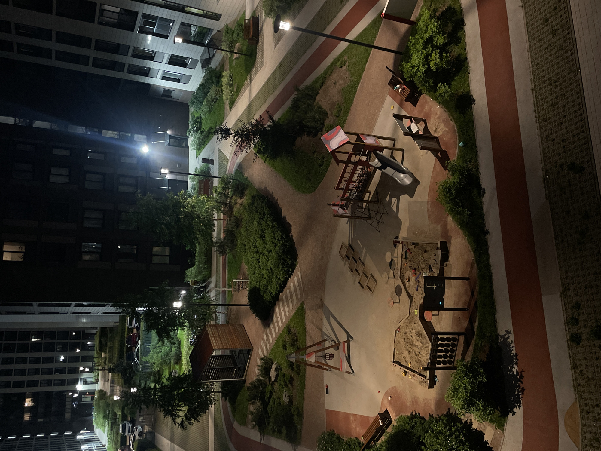 The image shows a nighttime view of a modern urban courtyard in Sinyavino, Russia, featuring a children's playground and surrounding residential buildings. The playground includes a sandpit with various toys, a slide structure, a swing set, and a set of wooden steps. There are also benches and walking paths throughout the courtyard. Several tall residential buildings with illuminated windows are visible, indicating the presence of inhabitants. Streetlights illuminate the area, casting shadows from trees and playground equipment. Cars are parked along the roadside in the background. The overall scene depicts a well-maintained, contemporary residential complex at night.