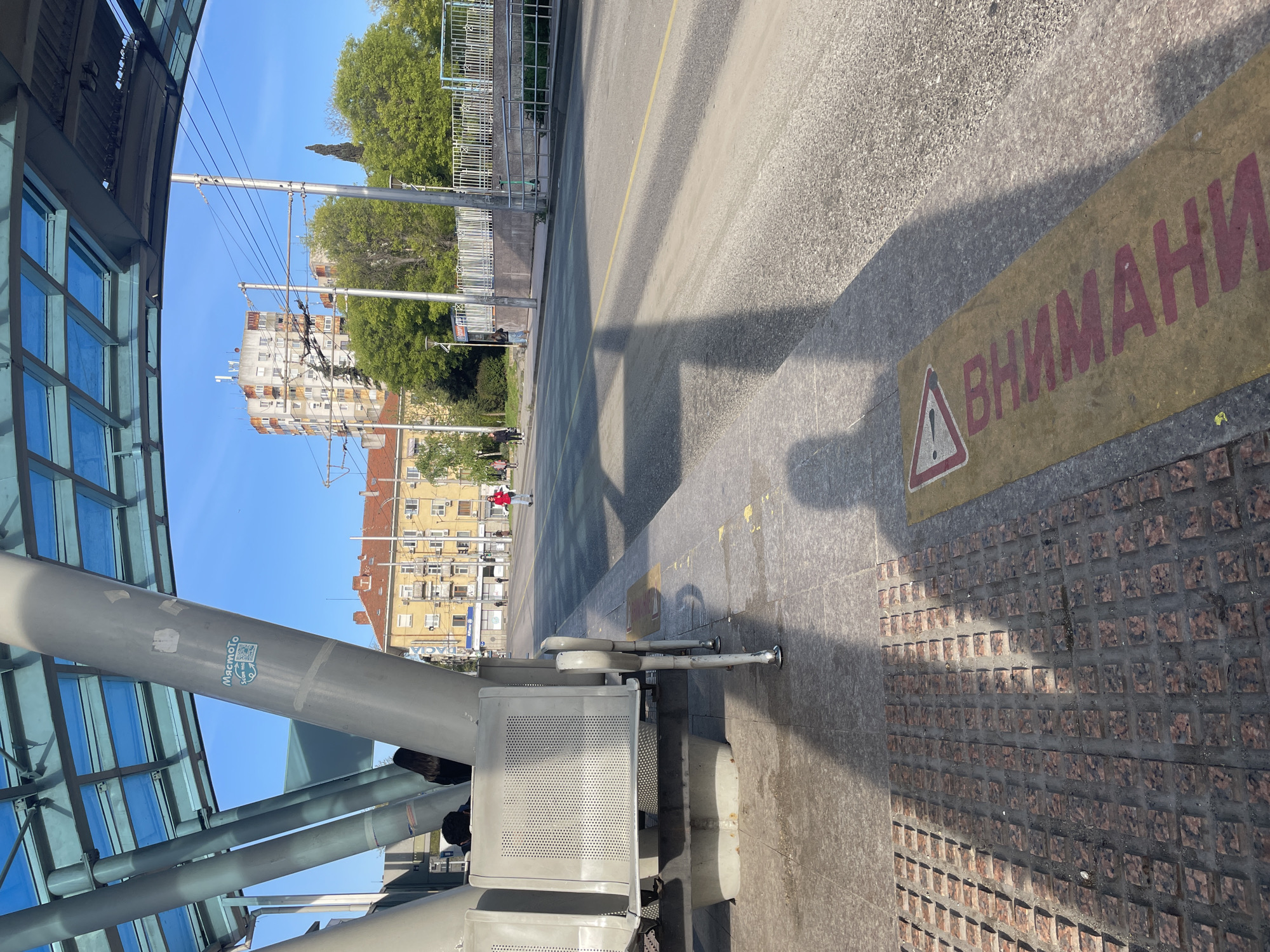 The image depicts an urban scene at a tram stop in Burgas, Bulgaria. The perspective is from within a modern tram station structure, characterized by a glass and steel frame design. The station has seating for travelers. Across the tram tracks, apartment buildings in yellow, white, and orange tones are visible. A pedestrian in red and white is walking along the sidewalk. A warning sign in Bulgarian, "ВНИМАНИЕ" (Attention), is placed next to the tram tracks. The ground is a mix of concrete and textured paving stones. The sky is bright blue, suggesting clear weather. Overhead tram power lines are present.