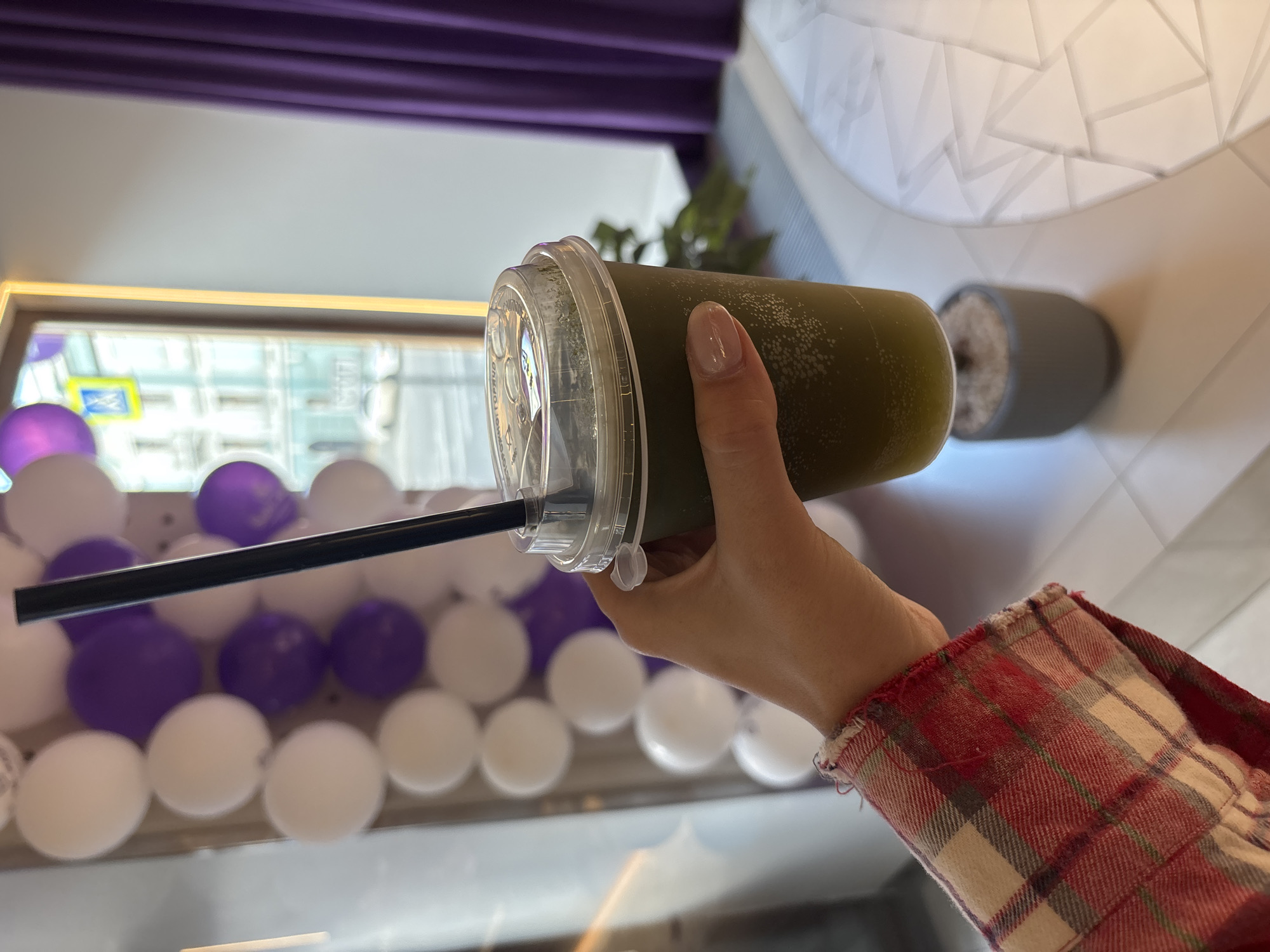 The image shows a person holding a green drink in a clear plastic cup with a black straw. The person is wearing a red and white plaid shirt. In the background, there's a window with a view of a building across the street. There are white and purple balloons visible near the window. The image was taken in Krasnoyarsk, Russia. A decorative element resembling a white geometric pattern is present to the right of the subject, suggesting an indoor setting.