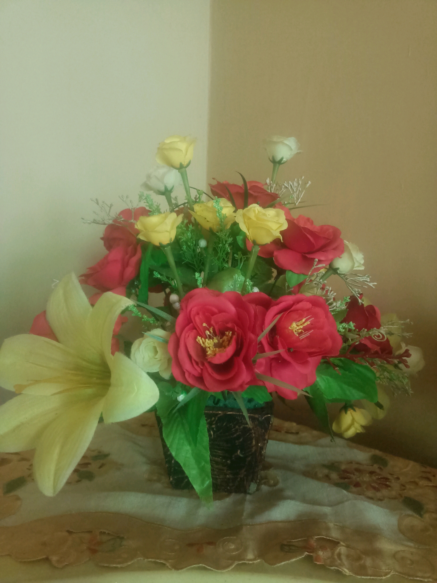 The image displays an artificial flower arrangement consisting of various roses in shades of red and yellow, along with a large, pale yellow lily. The flowers are presented in a dark, textured vase. Green artificial foliage is interspersed throughout the arrangement. The vase is placed on a surface covered with a patterned cloth that appears to have floral designs and a decorative, scalloped edge. The background consists of a wall that transitions from a lighter color on the left to a darker, warmer tone on the right. The lighting suggests an indoor setting. There are no people or discernible actions depicted in the image. The scene is static and focuses solely on the floral arrangement and its immediate surroundings. The location of Trenggalek, Indonesia, is not visually indicated within the image itself.