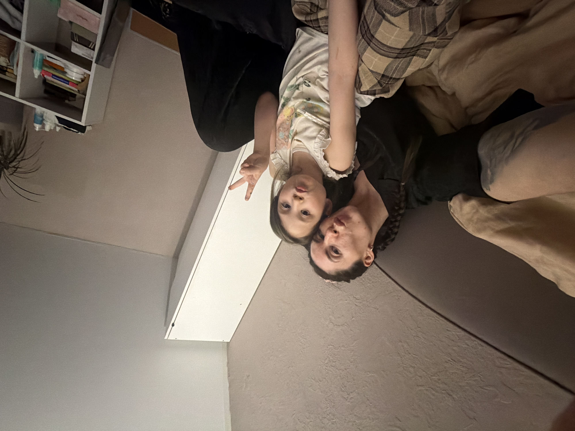 The image depicts an indoor scene, described as being in Perm, Russia, featuring an adult female and a juvenile female. The adult female, positioned on the left side of the frame, has dark hair styled in braids and wears a dark collared top. She looks directly towards the camera with pouted lips and is leaning against a dark, possibly upholstered, headboard or cushion. Adjacent to her on the right is a juvenile female, also with dark hair, wearing a light-colored sleeveless top with a subtle pattern and plaid shorts. She is holding her right hand in a peace sign gesture and has pouted lips, looking directly at the camera. Both individuals are seated on a surface covered by a beige duvet and a brown and beige plaid blanket. The background to the upper left features a light-colored, textured wall. To the right, a white vertical panel, likely part of a cabinet or storage unit, angles sharply upwards towards a plain light-colored ceiling. In the lower right, a white open shelving unit with multiple compartments holds various items, including books, bottles, and a potted plant with dark, spiky leaves. The scene is illuminated by artificial light.