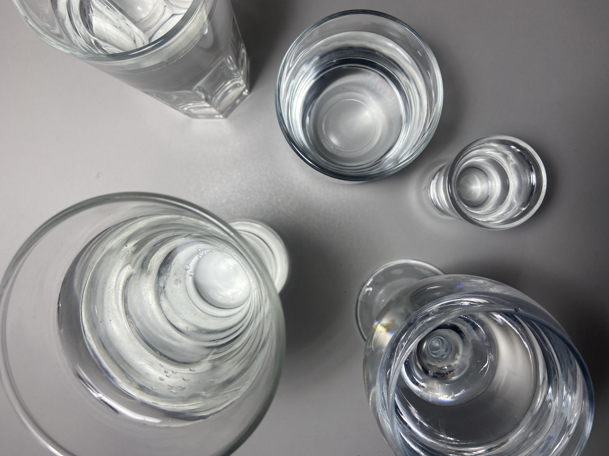 The image displays an overhead, top-down view of five clear glass vessels, each containing a transparent liquid, likely water, resting on a flat, light grey surface. No people are present, and no actions or interactions are depicted.
From left to right and top to bottom:
1. **Top left:** A large, cylindrical glass tumbler filled with liquid, exhibiting visible air bubbles near its interior base.
2. **Top right:** A hexagonal-sided glass tumbler, partially visible and also containing liquid.
3. **Bottom left:** A stemmed glass, possibly a wine glass or goblet, featuring a distinct swirling pattern in its design and filled with liquid. Subtle iridescent reflections are visible on its rim.
4. **Middle right:** A smaller, round glass, cylindrical in shape, filled with liquid.
5. **Bottom right:** The smallest glass, also round and cylindrical, filled with liquid.
The glasses are arranged somewhat haphazardly across the grey surface, which appears to be a plain tabletop or counter. The lighting is even, creating subtle reflections and shadows. The environment is sterile and lacks any distinguishing features or background elements that would indicate a specific location. The image contains no identifiable markers or contextual clues to determine a location within the city of Budva, Montenegro, or any other geographic area.