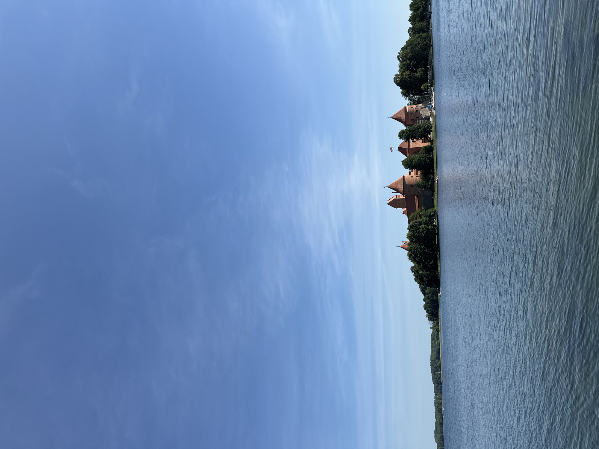 The image depicts Trakai Island Castle, a medieval castle located in Trakai, Lithuania. The castle is situated on an island in the Galvė Lake, surrounded by lush green trees. The sky is a clear, bright blue with a few wispy clouds. The water of the lake is calm and reflects the blue of the sky. Several boats are visible on the lake, and a flag flies from one of the castle towers. The architecture of the castle is characterized by its red brick walls and pointed towers, giving it a formidable and historic appearance. The scene is bathed in natural daylight, suggesting it was taken on a clear day, likely during the warmer months given the foliage.