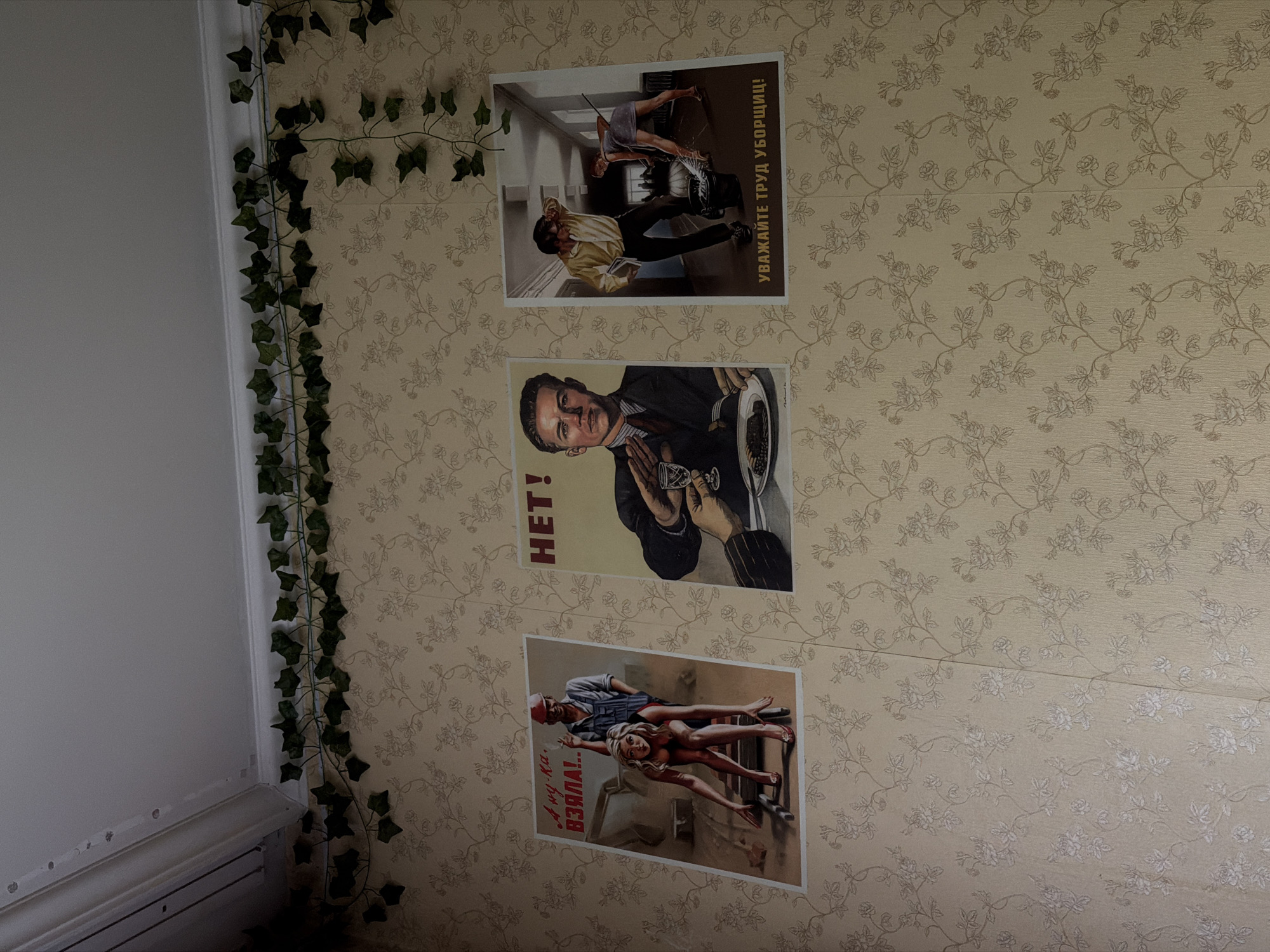 The image shows three Soviet-era propaganda posters hanging on a wall with a floral wallpaper pattern. The posters feature stylized illustrations and Russian text. The first poster, on the left, depicts a woman in a red dress and high heels, posed provocatively on a ladder, with a man in overalls and a red cap behind her, smoking a cigarette. The text above them reads "А ну-ка, взяла!!" (roughly translates to "Come on, grab it!"). The second poster, in the center, shows a stern-faced man in a suit, holding a glass of wine and a fork, with a plate of food in front of him. A large red exclamation mark is visible to the left of the man. The text above reads "НЕТ!" ("NO!"). The third poster, on the right, illustrates two people in a hallway. A man in a yellow shirt and black pants is carrying a book, while a woman in a dress is mopping the floor. The text below reads "УВАЖАЙТЕ ТРУД УБОРЩИЦ!" ("RESPECT THE WORK OF CLEANERS!"). A decorative green vine with artificial ivy leaves is draped along the top of the wall, near the ceiling molding. The lighting suggests an indoor setting, possibly a room or hallway. The location is not identifiable from the visual information provided.