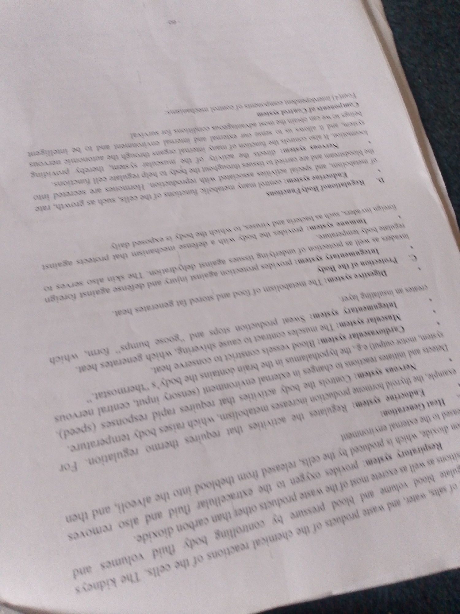 The image displays a close-up, angled view of a page from a textbook or educational material. The page is filled with densely packed text, primarily in English, describing various biological systems and their functions. The text is printed in a dark ink on white paper, which appears slightly yellowed and creased, suggesting it is an older document. The angled perspective and lighting create shadows and glare on the page, making some parts of the text difficult to read.
The visible text includes headings and descriptions for biological systems such as the "Nervous system," "Endocrine system," "Respiratory system," "Muscular system," "Integumentary system," "Digestive system," and "Immune system." Specific functions mentioned include controlling body activities, regulating body temperature, providing protection against injury and foreign invaders, and enabling survival through adaptation to the external environment. Some phrases hint at the complexity of these systems, like "control mechanisms" and "metabolic functions."
There are no people, objects, or any scene beyond the textbook page itself. The setting is implied to be an indoor environment where reading or studying would take place. No specific time of day, weather, or emotional cues are present. The context of "Jalingo, Nigeria" is provided externally and is not derivable from the image content.