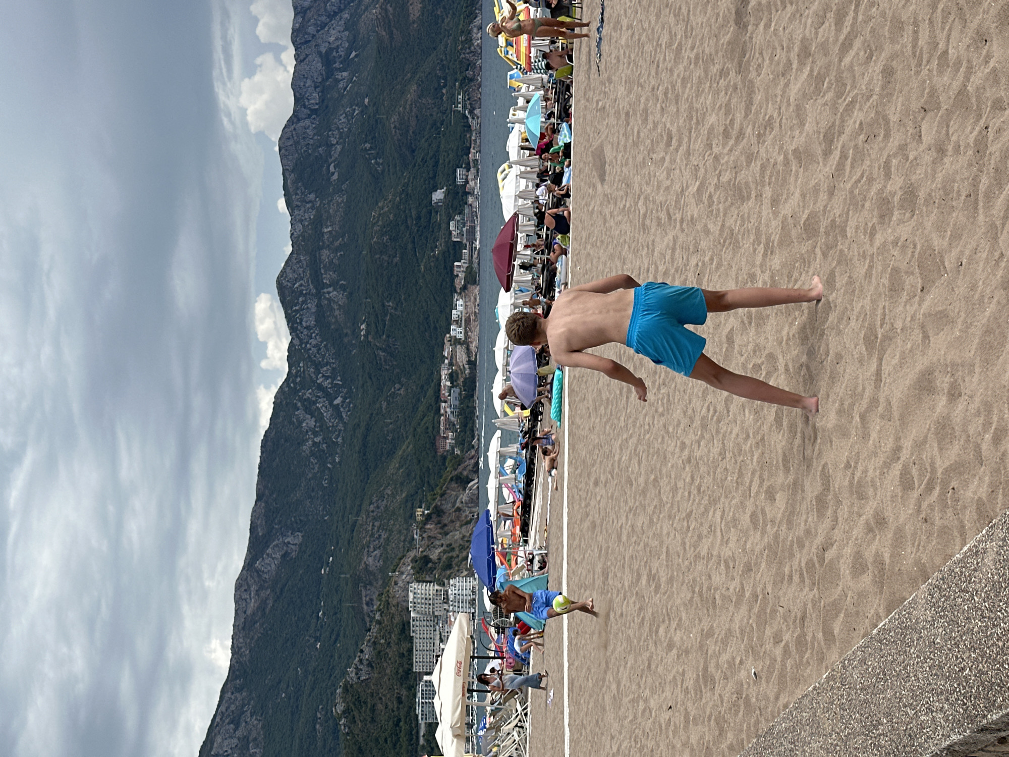 The image depicts a crowded beach scene in Boreti, Montenegro, with a backdrop of lush green mountains and buildings. In the foreground, two young boys are playing soccer on the sandy beach. One boy, wearing blue shorts, is in mid-kick, his body contorted as he strikes a yellow soccer ball. The other boy, partially visible on the right side of the frame, appears to be reacting to the play. The beach is dotted with numerous beach chairs and umbrellas, indicating a busy day with many people enjoying the sun and sea. In the distance, the calm blue water of the Adriatic Sea is visible, with boats anchored near the shore. The sky is overcast with dramatic clouds, suggesting a possibly stormy or moody atmosphere, contrasting with the lively beach activity.