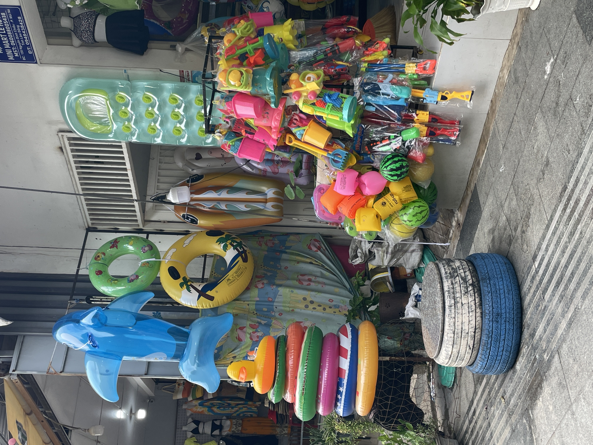 This scene captures an outdoor retail display in Phu Thuy Ward, Vietnam. Dominating the foreground are numerous inflatable items: a large, blue dolphin, several colorful inner tubes of varying diameters, and a green inner tube. Further back, there are inflatable surfboards and a large, light blue inflatable air mattress. On the right side are stacks of toys, water guns and plastic beach toys. Beside this, two painted tires serve as makeshift furniture, stacked upon each other. The photograph is taken on a sidewalk along a street. A sign indicating "MINI MART" with contact information in Vietnamese is partially visible at the top left of the image.
