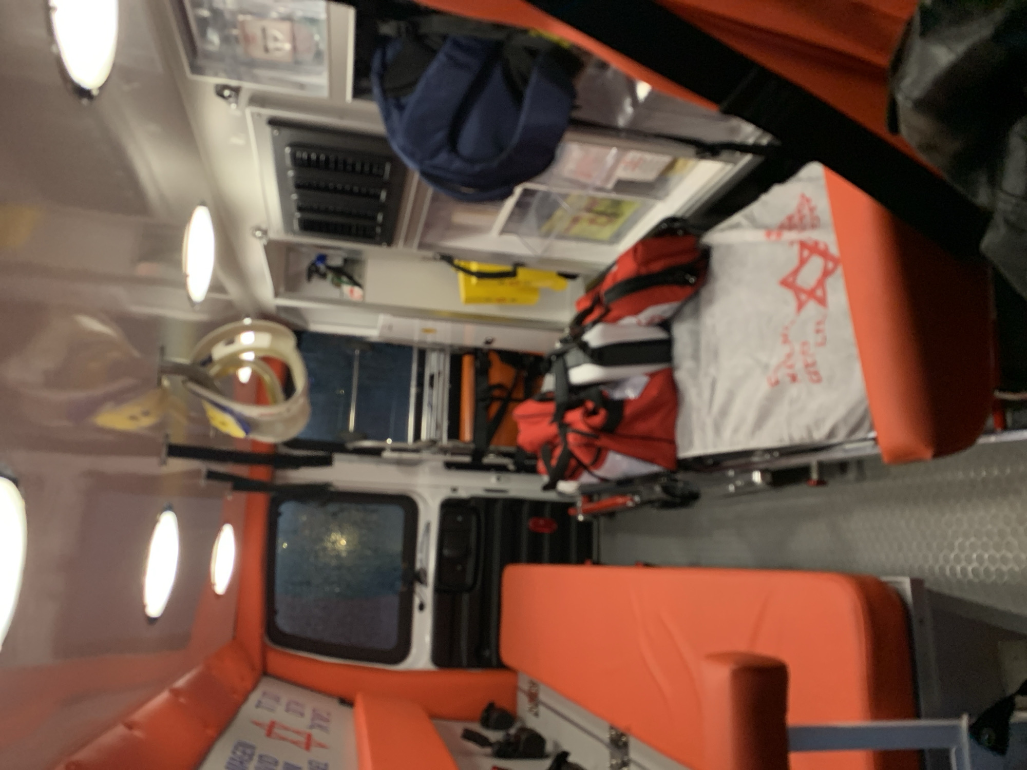 The image shows the interior of an ambulance. The walls and ceiling are white with built-in oval lights. Orange padding lines the lower walls and seating areas. There is a window on the left side of the frame. Medical equipment and supplies are stored in compartments along the upper part of the ambulance. A stretcher with medical bags and a white cloth featuring a Star of David pattern is visible. The location is presumed to be Tel-Aviv, Israel, based on the Star of David.