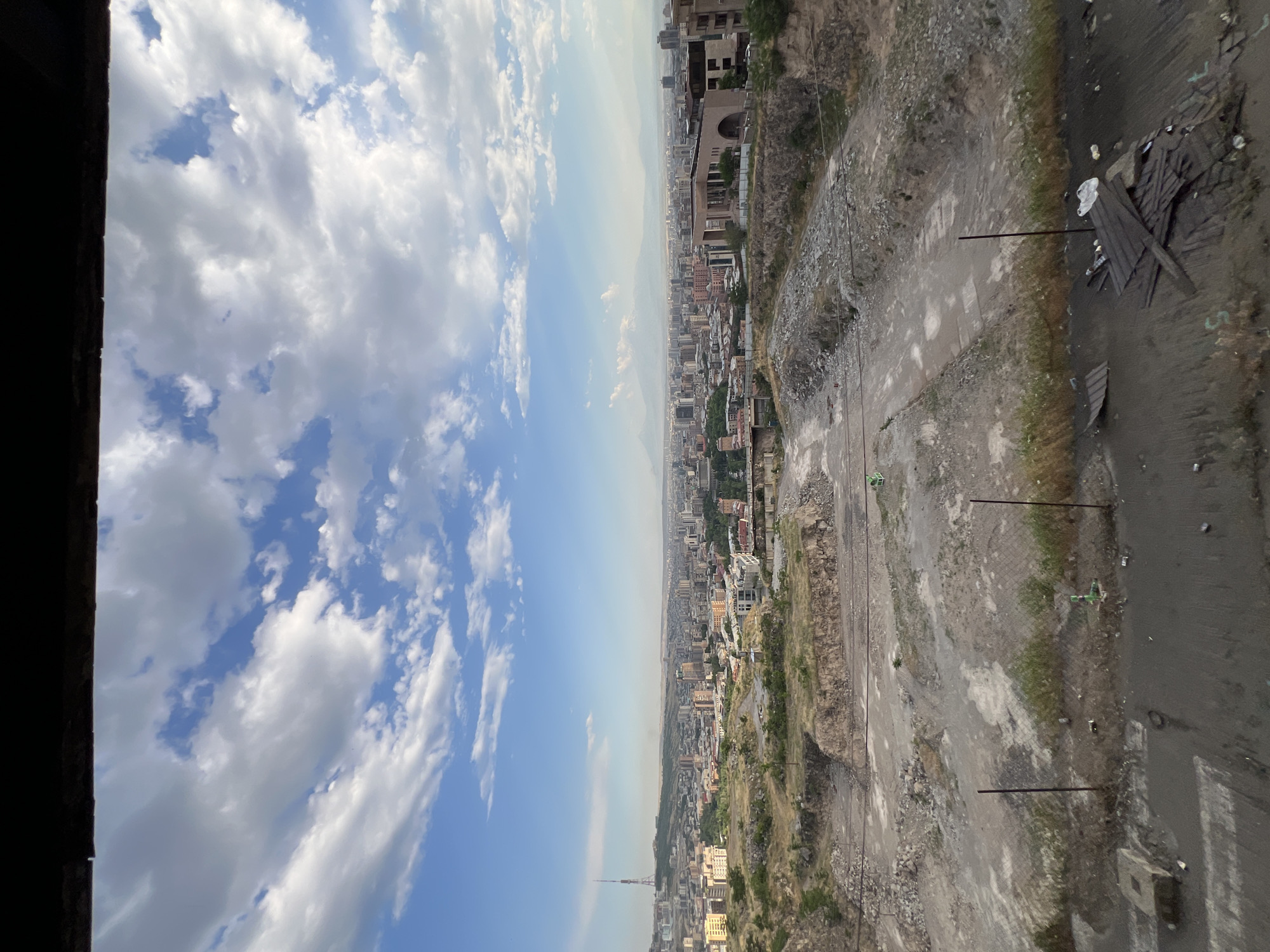 This is an aerial, wide-angle, outdoor, daytime shot of a panoramic view of Yerevan, Armenia, taken from an elevated position. The foreground is dominated by a dry, dusty, barren landscape with rocky outcrops, dirt paths, and patches of dry grass. A wire fence runs horizontally across the middle ground, with scattered debris, including wooden planks, plastic bags, and cans, visible on the ground. Beyond the barren foreground, the middle ground transitions into a dense urban cityscape with numerous buildings of varying heights and architectural styles. In the distance, more buildings extend towards the horizon, punctuated by a prominent radio tower on a hill to the left. The sky above is a vibrant blue, filled with fluffy white and grey clouds, suggesting fair weather. In the far distance, faint outlines of mountains are visible, possibly Mount Ararat. The overall scene depicts a contrast between a neglected, undeveloped area in the foreground and the bustling city beyond, with a natural mountain landscape in the background.
