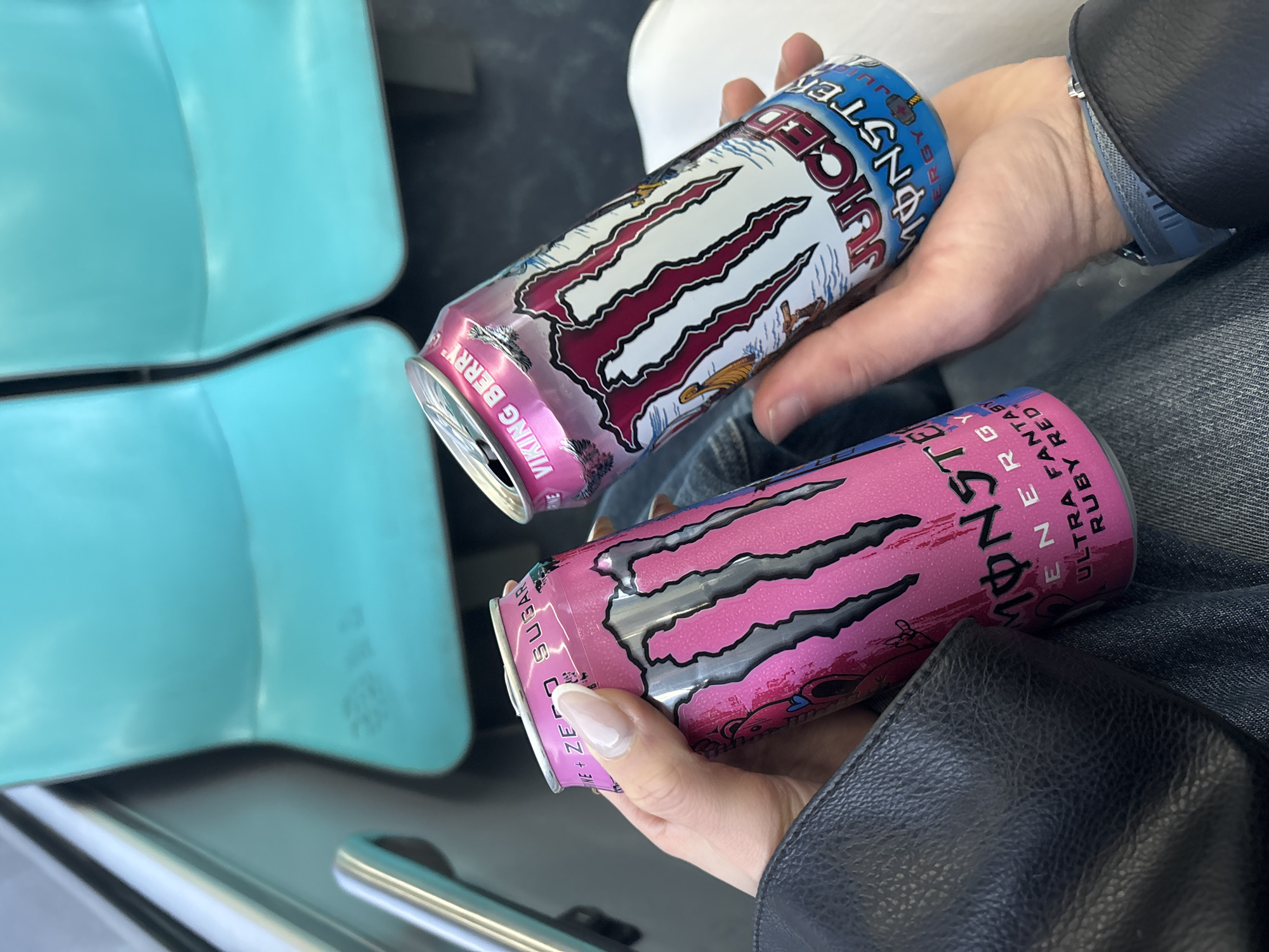 The image captures a close-up of two hands holding two Monster energy drink cans within what appears to be a vehicle or public transport setting. In the background, two light blue, possibly plastic or upholstered, seats are visible, along with a metal railing in the lower foreground.
One hand, with neatly manicured nails, holds a pink Monster Energy Ultra can. This can features a prominent black Monster claw logo and is identified by the text "MONSTER ENERGY ULTRA FANTASY RUBY RED" and "ZERO SUGAR".
The other hand holds a white, blue, and pink Monster Juiced energy drink can. This can also displays the iconic black Monster claw logo outlined in white and red, with the text "JUICED MONSTER ENERGY" and the flavor "VIKING BERRY".
Partial views of a black leather jacket sleeve and blue denim jeans suggest the person's attire. A wristwatch is also visible on one wrist. The image focuses sharply on the hands and cans, with the background providing a slightly blurred context of an interior space, likely in transit.
