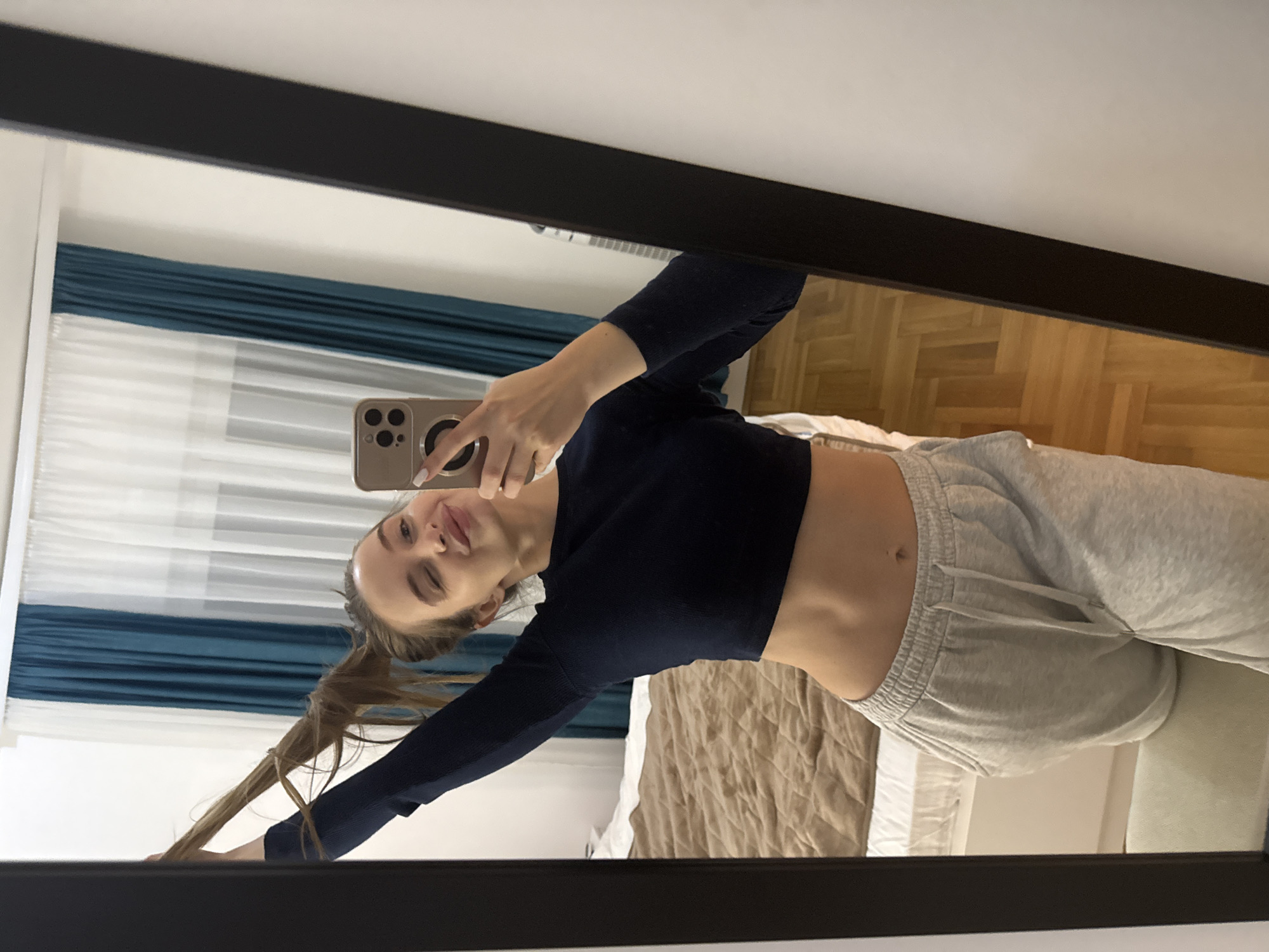 A young woman is reflected in a mirror, taking a selfie with a phone. She has light brown hair pulled into a ponytail and is wearing a dark blue long-sleeved crop top and light gray sweatpants. The background visible in the mirror shows a room with hardwood floors, a bed with a beige duvet, and blue and white curtains. The photo was likely taken indoors, possibly in a bedroom or apartment. It is not possible to pinpoint the exact location in Budva, Montenegro, as the image only shows an interior space.
