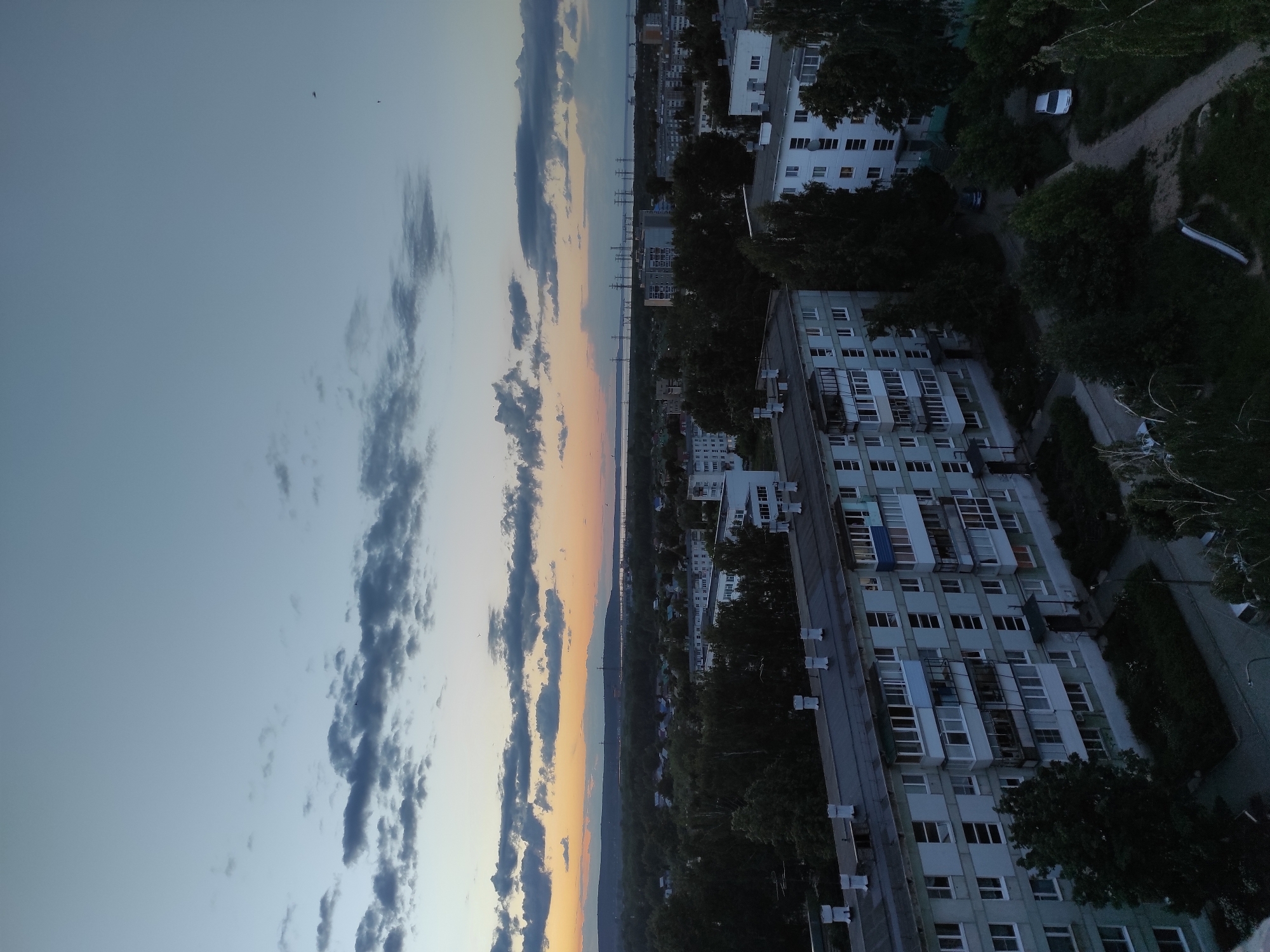 The image displays a high-angle view of a cityscape at dusk or dawn, oriented horizontally despite the original file's vertical rotation. The sky occupies the upper portion, showing a gradient from a light blue at the top to warm orange and yellow hues near the horizon, indicating either a sunset or sunrise. Scattered clouds, some elongated and dark, are visible across the sky. Several small, dark specks, possibly birds or distant aircraft, dot the sky.
In the middle ground, a wide body of water, likely a river or large lake, stretches across the horizon, with distinct silhouettes of power lines or transmission towers crossing it in the distance. The landscape beyond the water appears flat, suggesting a distant plain or low hills.
The foreground and mid-ground are dominated by multi-story residential buildings, likely apartment blocks, characterized by light-colored facades and numerous windows, many with enclosed balconies. Dense green trees and bushes are interspersed among the buildings, along with visible asphalt roads and dirt paths. A few cars are parked or moving on these paths, including a white car and a darker vehicle. The overall lighting is muted and soft, characteristic of twilight. There is no visible text in the image. The scene conveys a sense of peaceful urban life as the day transitions.