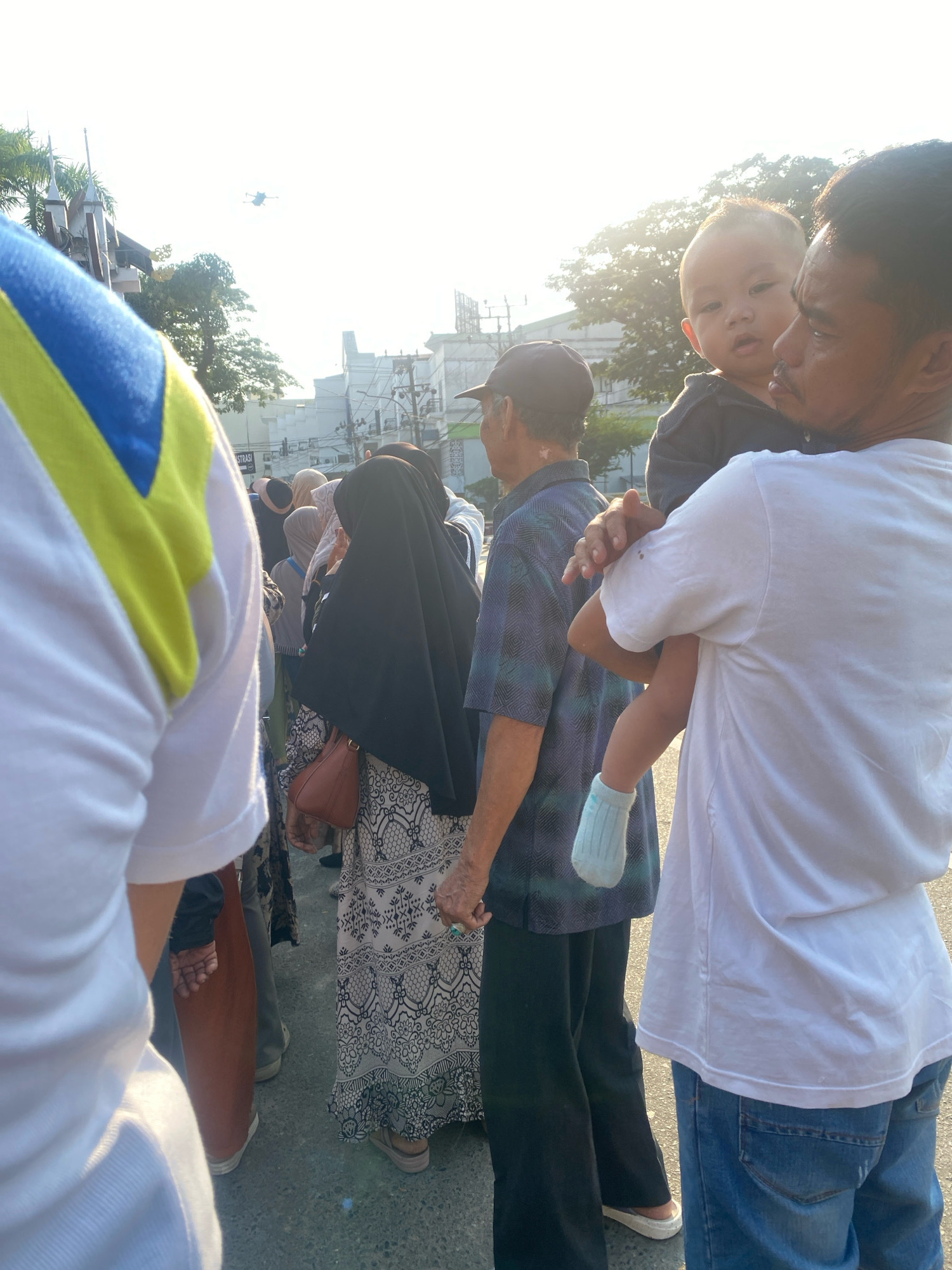 In Banjarmasin, Indonesia, a man in a white t-shirt holds a young child. The child, wearing a light blue sock on its right foot, looks towards the camera. Behind them, an elderly man in a patterned shirt and a black cap stands with his back to the viewer. To the left, a person in a white shirt with blue and yellow accents is visible, and further left, a woman in a black hijab and a patterned dress is seen. The scene appears to be outdoors on a paved surface, with trees and buildings in the background. The sky is bright, suggesting daylight, and a drone is visible in the sky.