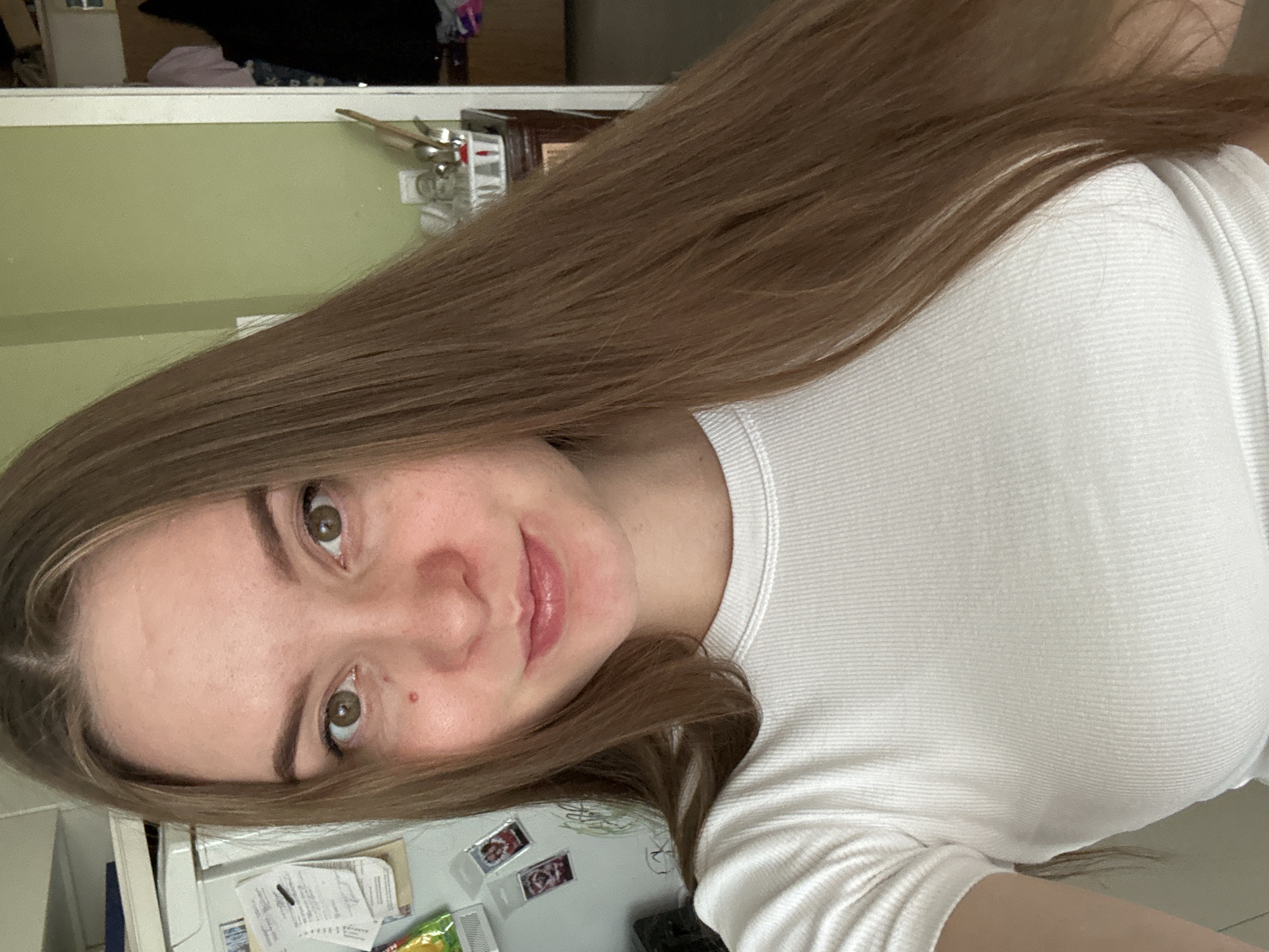 A young woman with long, brown hair is taking a selfie. She is wearing a white, ribbed t-shirt. Her skin is fair, with a few freckles and a small mole on her cheek. Her eyes are brown, and she has a slight smile. The background appears to be a kitchen area, with a refrigerator on the left and a green wall on the right. There are various items visible on the refrigerator and in the background, suggesting a domestic setting. The lighting is natural, coming from the front. The image does not provide any specific geographical clues that would indicate the location in the city of Noviy, Russia.