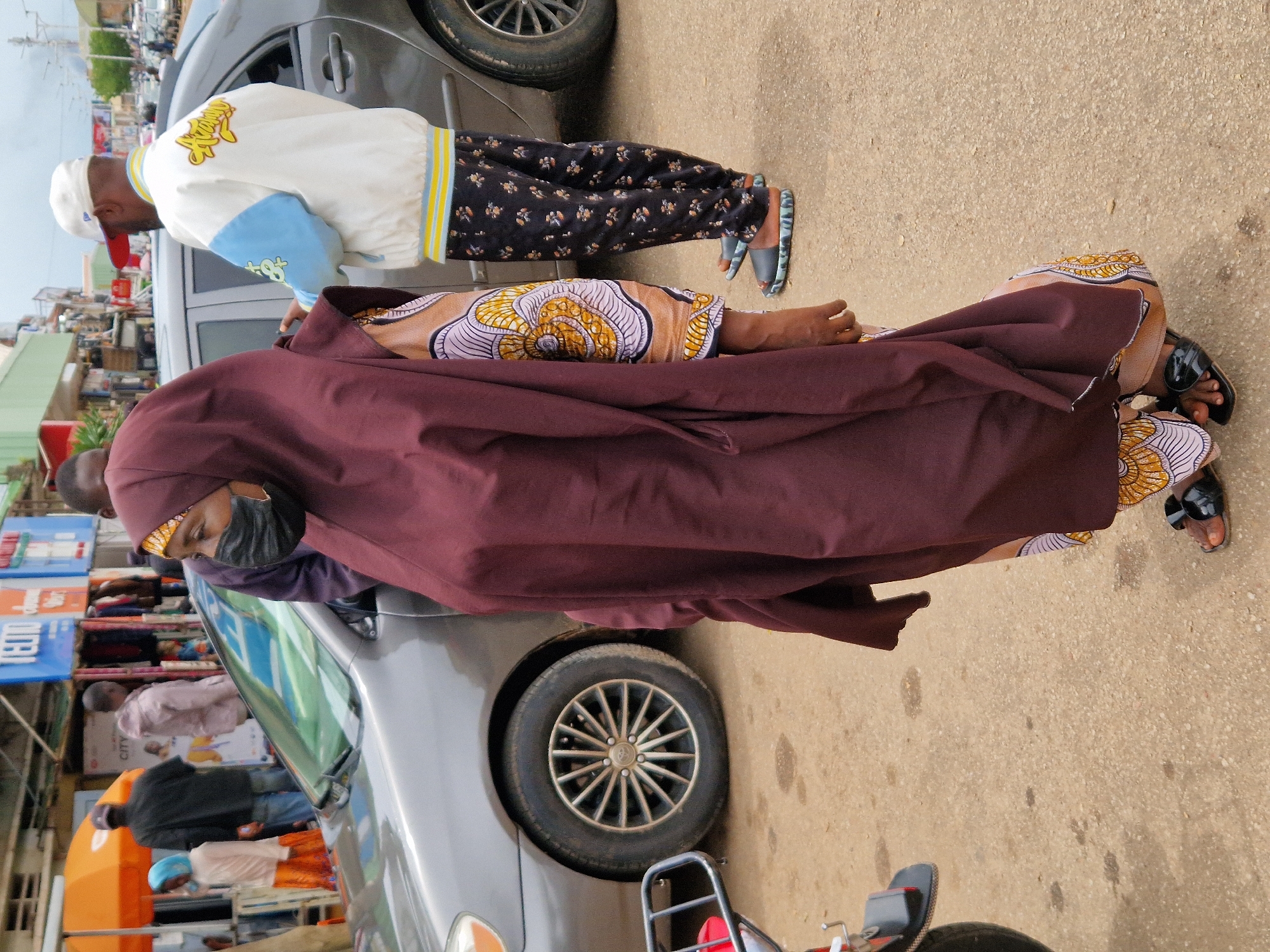 In an outdoor, urban environment, a group of people stand near a parked car. The primary subject is a woman wearing a dark maroon hijab and a patterned dress, her face partially obscured by a black face mask. She stands on a dirt or gravel road. To her right, a man in a white and blue bomber jacket with yellow trim and a white baseball cap is visible. He wears dark pants with a floral pattern. Behind them, the side of a grey sedan is prominent, with its front wheel showing. Further in the background, other individuals and a building with storefronts suggest a marketplace or commercial area. An orange umbrella and a motorcycle with a red component are also present in the scene. The lighting suggests an overcast day.