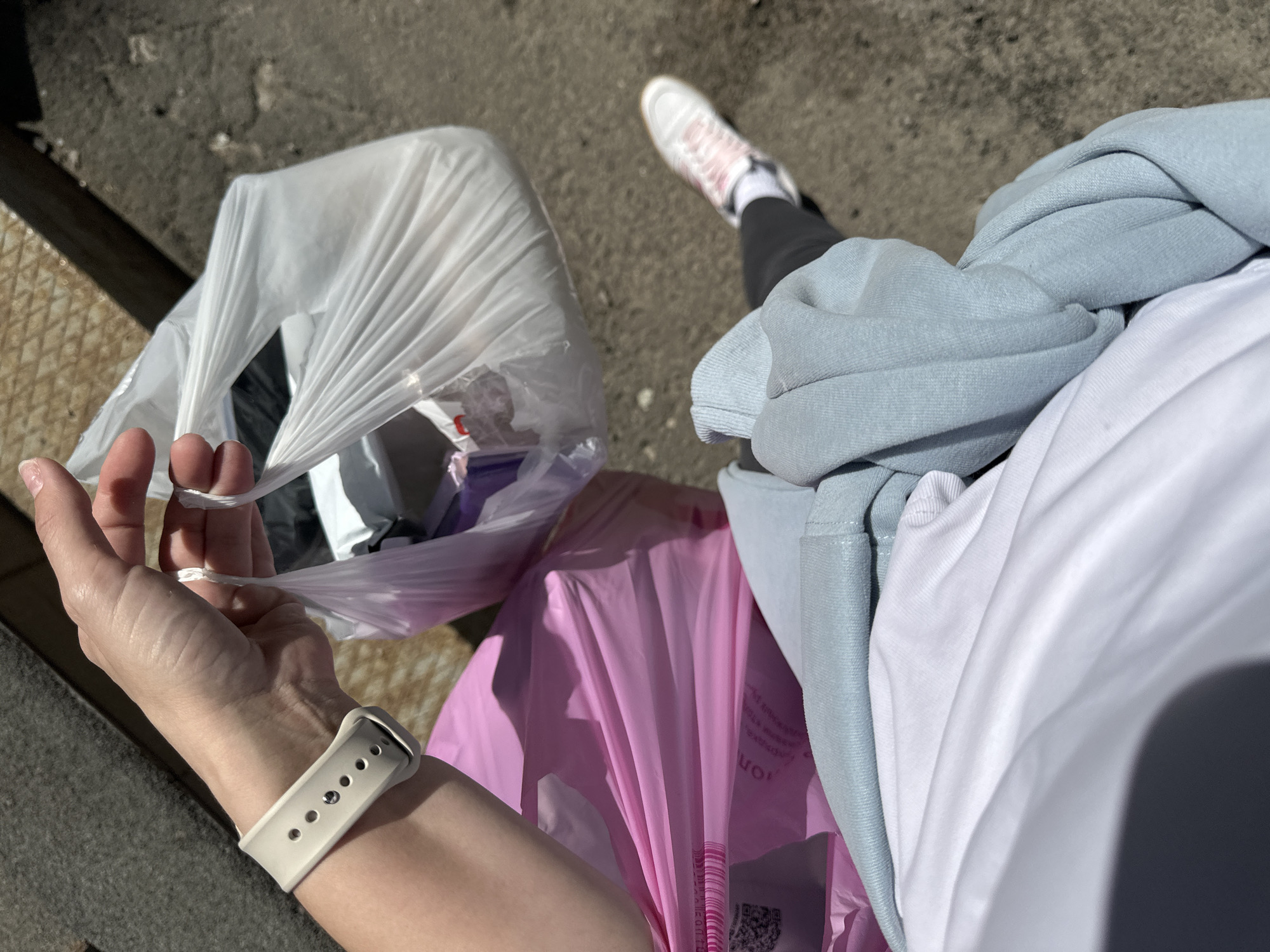 A first-person perspective photograph shows a person holding two shopping bags, one white and one pink, along with a light blue sweatshirt. The person is wearing a white t-shirt, black leggings, and white sneakers with pink accents. A smartwatch with a white band is visible on the person's wrist. The setting appears to be an urban environment, possibly in Perm, Russia, given the prompt's instructions, with a concrete surface and a metal grate partially visible in the background. The image shows the person seemingly in the act of carrying shopping bags and a sweatshirt while walking outdoors.