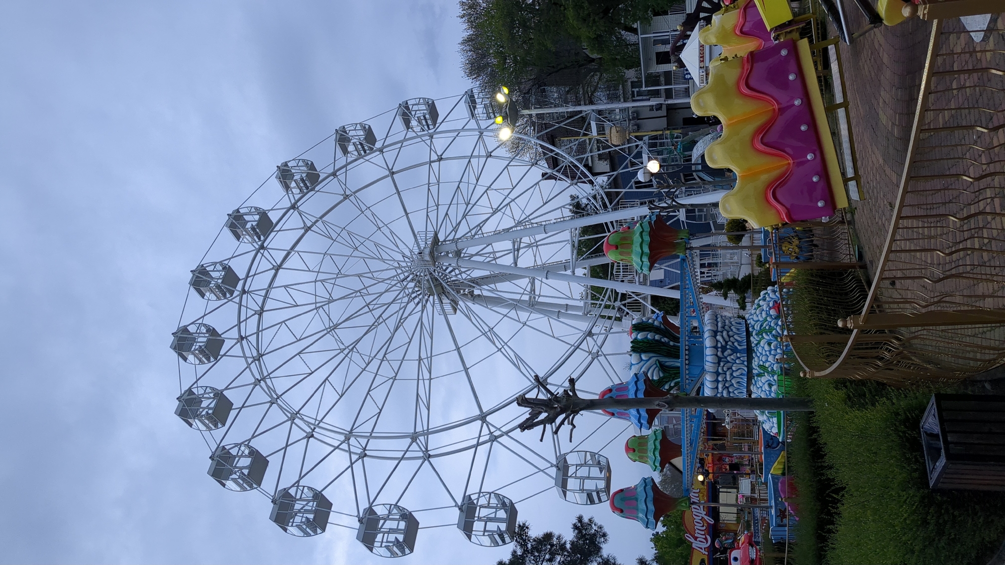 The image displays an amusement park scene in Gelendzhik, Russia, dominated by a large white Ferris wheel under an overcast sky. The Ferris wheel occupies the left and central parts of the frame, featuring numerous gondolas that appear empty.
To the right of the Ferris wheel, various colorful amusement park elements are visible. These include decorative structures resembling large, vibrant ice cream cones or cupcakes, and a multi-tiered blue and white feature designed to look like waves. Further right, a bright yellow and purple ride piece, shaped like a giant cake slice, stands alongside a golden, ornate metal fence. A paved area with a brick pattern is visible, and green bushes and trees are scattered throughout the park grounds. A dark wooden trash can is also present.
The sky is a uniform grey, indicating an overcast day or twilight hours. Some artificial lights on the park structures and in the background buildings are illuminated, reinforcing the impression of dusk or low light conditions. In the background, a building has the Russian word "КАССА" (CASH DESK) written on it, identifying it as a ticket office. Another ride structure features the partially visible Russian text "АВТОГОРОД" (AUTOCITY) along with a character resembling Lightning McQueen, suggesting a car-themed attraction. No people are visible, lending a quiet, expectant mood to the scene.