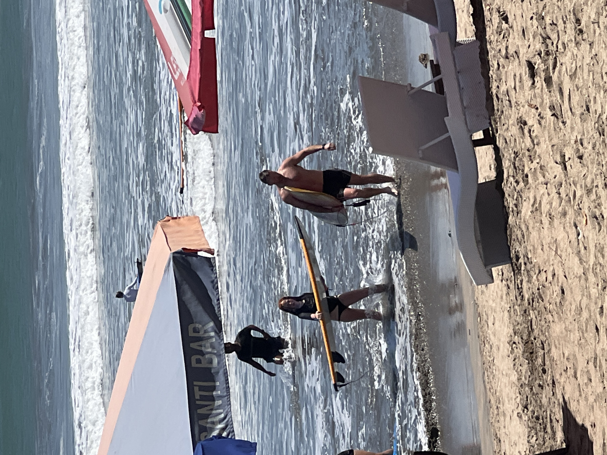 In Kuta, Indonesia, on a sunny day, two individuals are walking in the shallow water near the shore, both carrying surfboards. To the left, a woman with red hair, wearing a black bikini, is holding a surfboard horizontally. Behind her, a man in a dark t-shirt and shorts is wading through the water. To the right, a man with dark hair, shirtless and wearing black shorts, walks with his surfboard tucked under his arm. The ocean has moderate waves. In the foreground, beach chairs are partially visible on the sandy shore, and a large umbrella with the text "SANTI BAR" on it extends over the water. Another umbrella, predominantly red, is visible further back on the right.