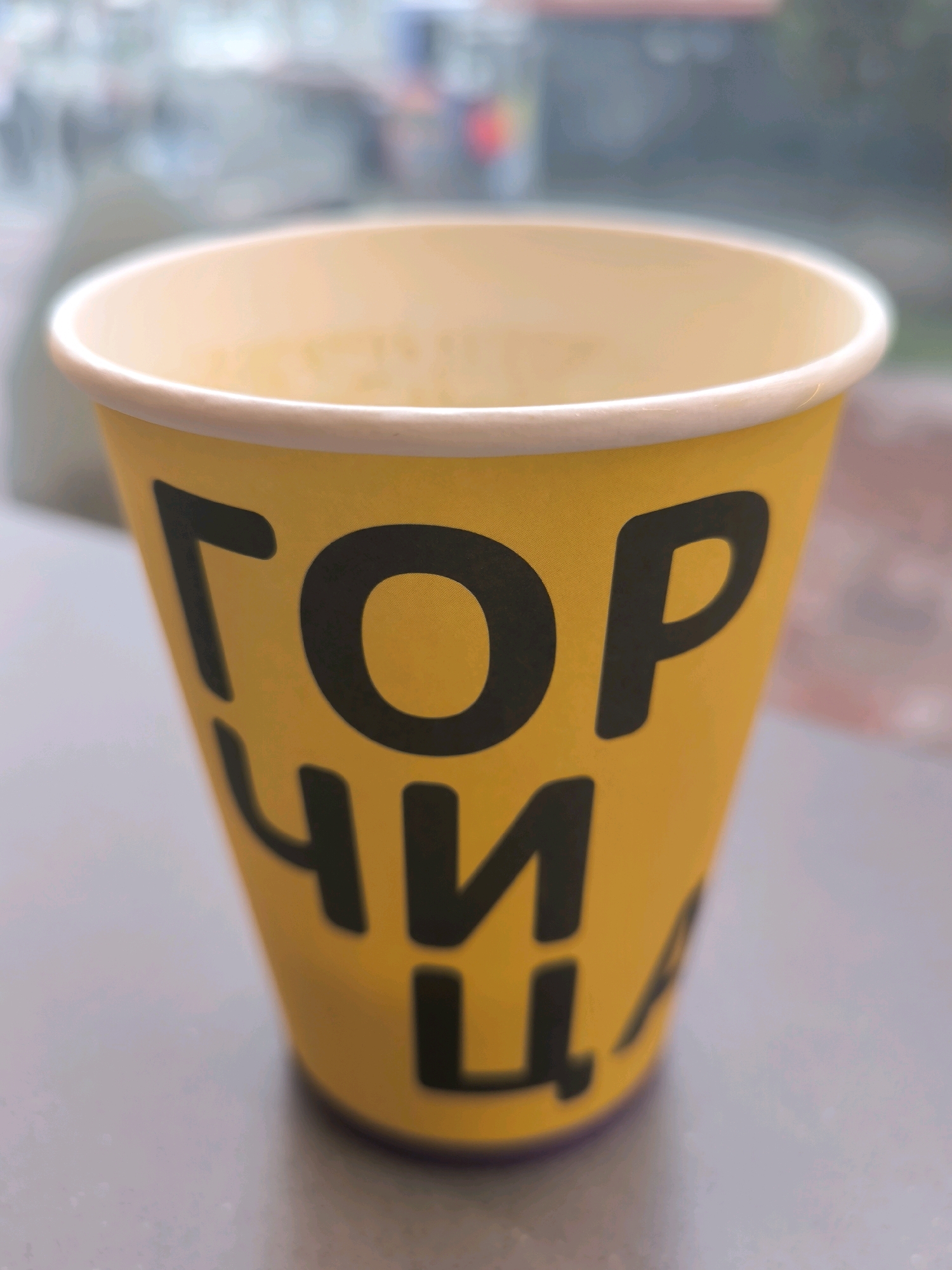 A close-up, eye-level shot shows a yellow paper cup with black text on a blurred background. The text on the cup, which appears to be in Cyrillic, reads "ГОРЧИЦА". The cup is slightly tilted, and the opening is visible at the top. The background is out of focus, showing shapes and colors that suggest an urban environment, possibly a street or cafe setting in Novosibirsk, Russia. The lighting is even, and there are no people or animals visible in the frame.