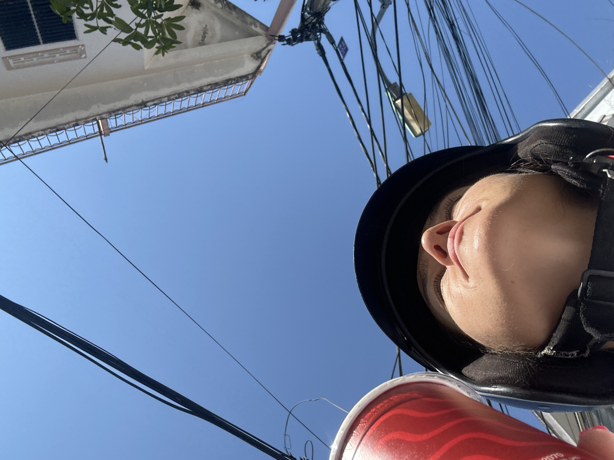 A low-angle shot depicts a person wearing a black helmet with a fastened strap. A red and white patterned disposable cup is visible in the foreground. In the background, the blue sky is crisscrossed by numerous power lines. Part of a building with a mesh railing and a window with closed shutters is on the left of the image and a light fixture is towards the center. The person in the helmet has fair skin, a closed mouth, and their eyes are closed. Based on the metadata, this photograph was taken in Bac Nha Trang Ward, Vietnam.