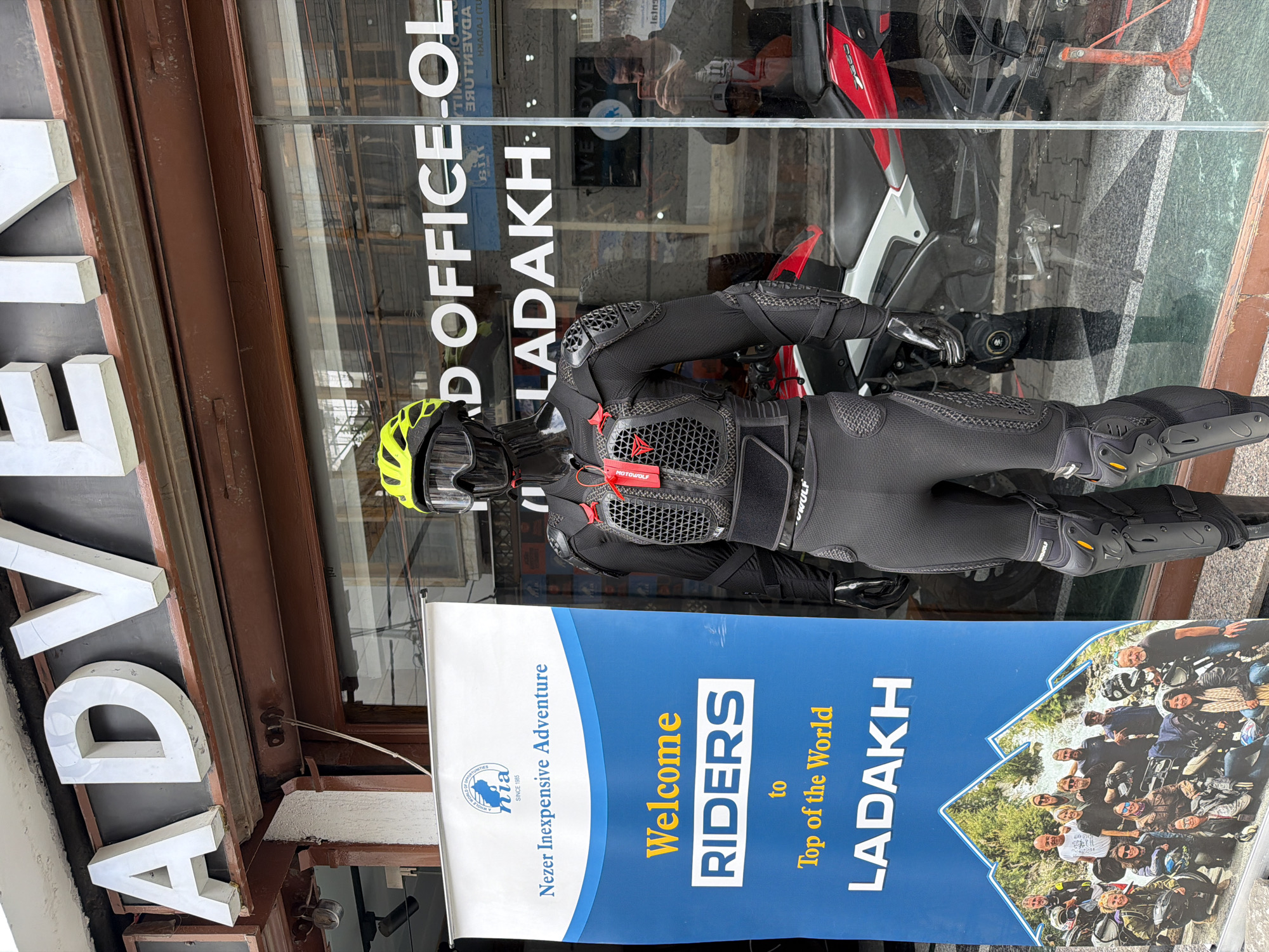 An outdoor shot captures a storefront in Leh, India, featuring a mannequin dressed in motorcycle protective gear. The mannequin is positioned in front of a large blue banner that reads "Welcome RIDERS to Top of the World LADAKH." Behind the mannequin and the banner, a large glass window displays the store's interior, with white lettering on the glass indicating "AD Office" and "LADAKH". The reflection in the window shows a man holding up a phone, likely taking a picture. A red and white motorcycle is visible through the glass, partially behind the mannequin. The storefront is adorned with large, metallic "ADVENTURE" lettering across the top. The environment appears to be a commercial street in Leh, characterized by traditional architecture and a clear daytime sky.