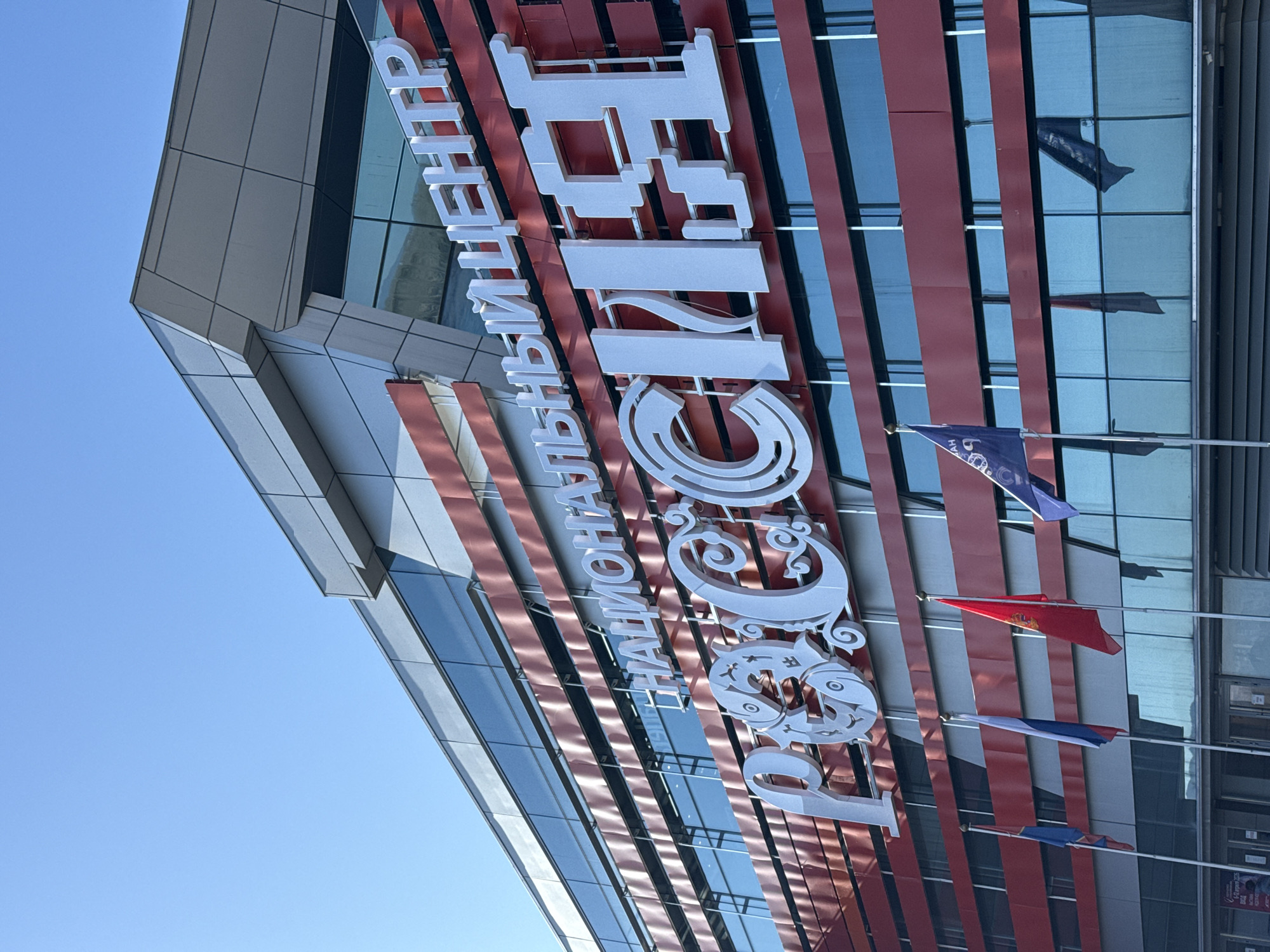 A low-angle, eye-level shot of the exterior of the "Rossiya – Moya Istoriya" (Russia – My History) historical park in Krasnoyarsk, Russia, showcases the modern building with its geometric facade. The building features a predominantly glass exterior with vertical red and gray stripes. Large, white Cyrillic letters spelling out the name of the park are mounted on the facade. A row of flags, including the Russian flag, is visible in front of the building. The sky is clear and blue.