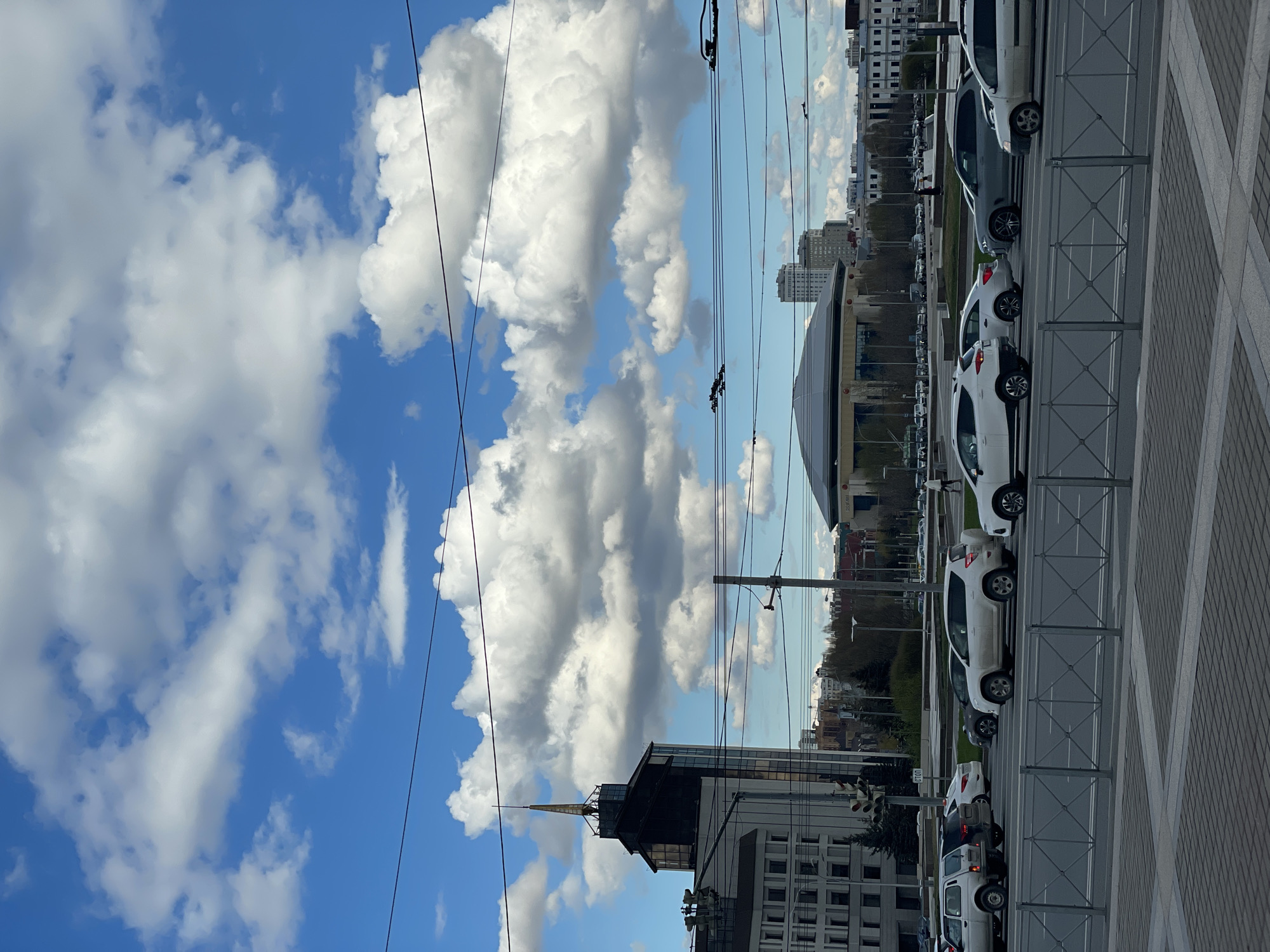 The image presents a vibrant urban street scene in Kazan, Russia, under a bright blue sky adorned with large, fluffy white and grey cumulus clouds. The view looks along a city street lined with buildings and parked vehicles.
In the foreground and mid-ground on the right, a row of predominantly white and light-colored cars are parked parallel to the road. They are separated from a grey, geometrically patterned paved sidewalk by a grey metal barrier fence. The road itself is visible beyond the fence.
The cityscape extends into the background with various multi-story buildings. A distinctive structure with a dark, pointed spire or roof element is visible on the left side. Further down the street, a large, dark, dome-shaped building, possibly a stadium or arena, stands out against the horizon. Overhead, numerous black power lines or tram wires crisscross the sky.
On the left side of the street, near a traffic light pole, a white 'STOP' sign is clearly visible. The bright daylight indicates it is daytime, likely morning or afternoon, with clear weather despite the significant cloud cover. The overall scene depicts a typical city environment on a pleasant, cloudy day.