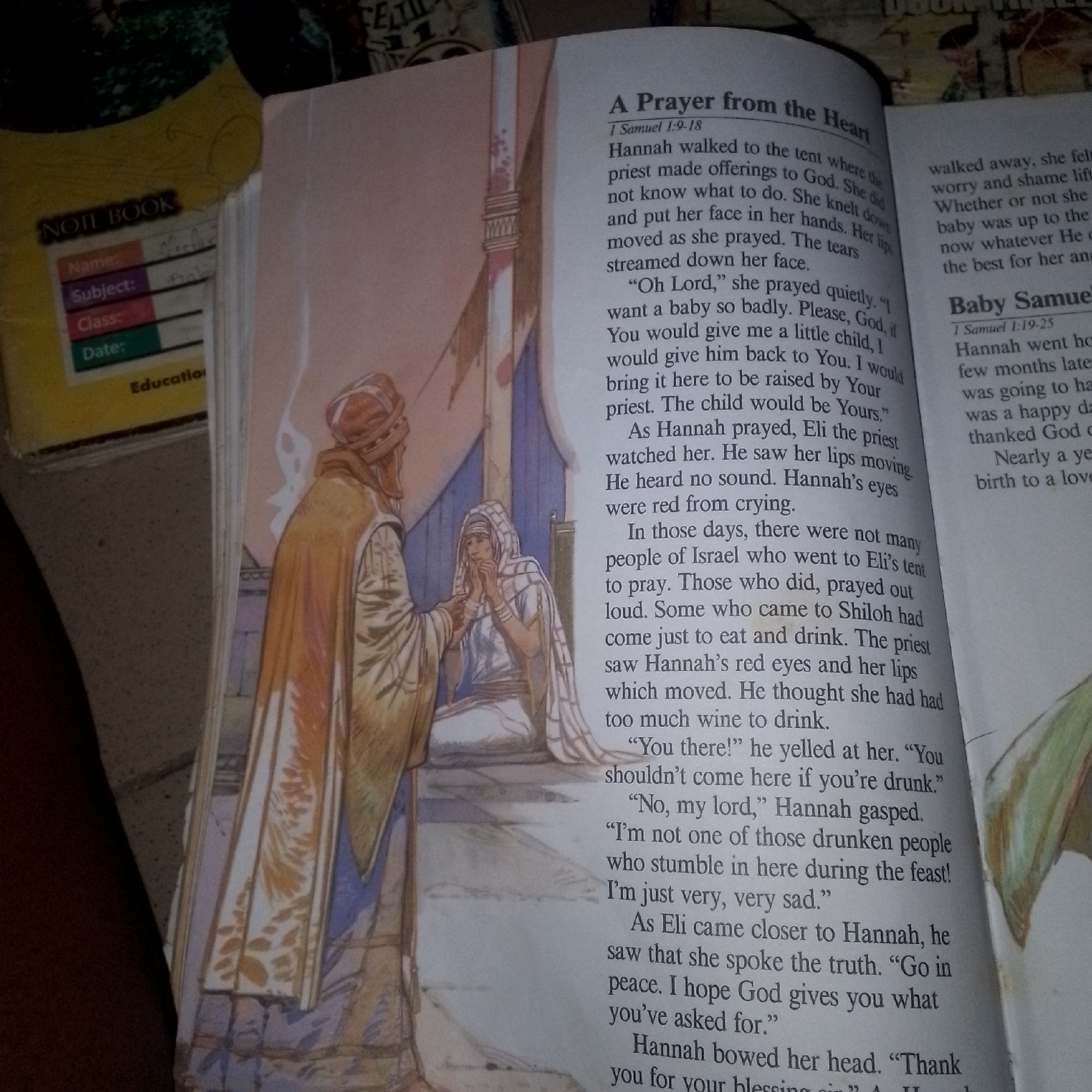 An open book displays a watercolor illustration on the left page and printed text on the right. The illustration depicts two figures: a man on the left wearing a turban and a long, flowing robe, and a woman on the right wearing a headscarf and a similar robe. The woman appears to be praying or crying, with her hands clasped to her face. The man stands to her left, seemingly observing her. The background of the illustration shows architectural elements suggestive of a tent or building.
The right page contains two sections of text. The top section is titled "A Prayer from the Heart" and references "1 Samuel 1:9-18." The text recounts Hannah's prayer and her interaction with Eli the priest. Below this is another section titled "Baby Samuel" referencing "1 Samuel 1:19-25," which continues the narrative about Hannah and her child.
In the upper left corner of the image, a portion of a yellow notebook with handwritten labels like "Name," "Subject," "Class," and "Date" is visible, along with the word "NOTE BOOK." The overall setting appears to be indoors, with the book laid flat on a surface.