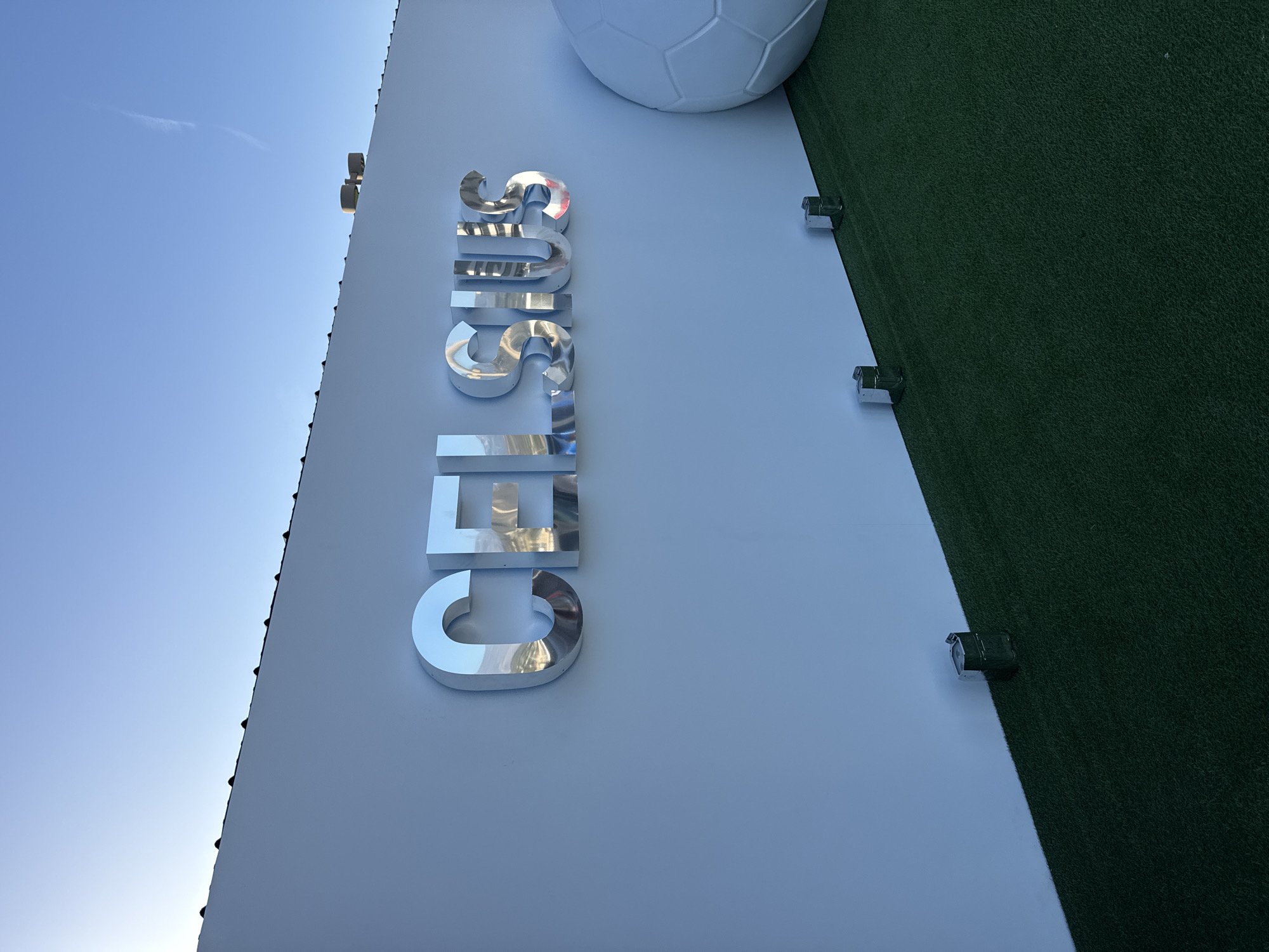 The image shows a sideways view of the word "CELSIUS" in large, chrome block letters mounted on a white wall. A partial soccer ball is visible at the top right corner. Adjacent to the wall, to the right, is a strip of green artificial turf. The photo is taken from below, looking upward. The sky is visible in the upper left corner, suggesting an outdoor location. The presence of the "CELSIUS" branding alongside a soccer ball and artificial turf suggests this is a promotional display, most likely in Glendale, United States of America, given it is a primary location for Celsius branding.