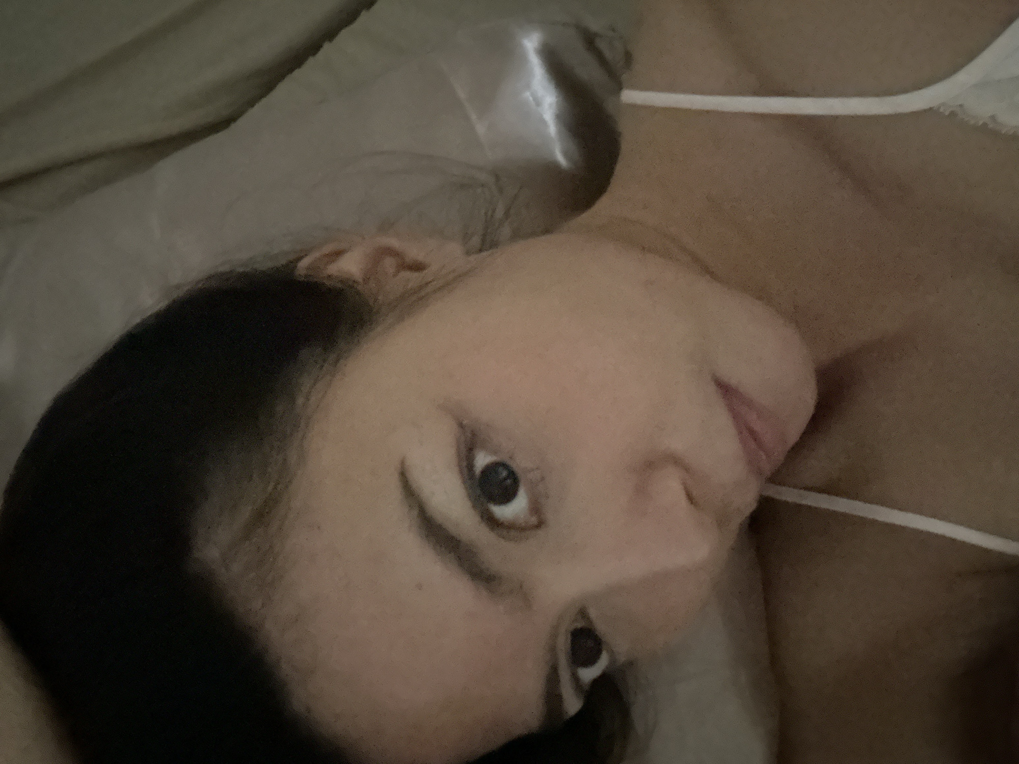 A close-up, overhead shot shows a woman lying down, with her head resting on a pillow. She has dark hair pulled back and is looking directly at the camera with wide eyes. Her face is illuminated from the front, highlighting her features and a few freckles. She is wearing a white spaghetti strap top. The background consists of a pillow and bedding, appearing to be indoors. No specific location in Khlong Luang Town Municipality, Thailand, can be identified from the image.