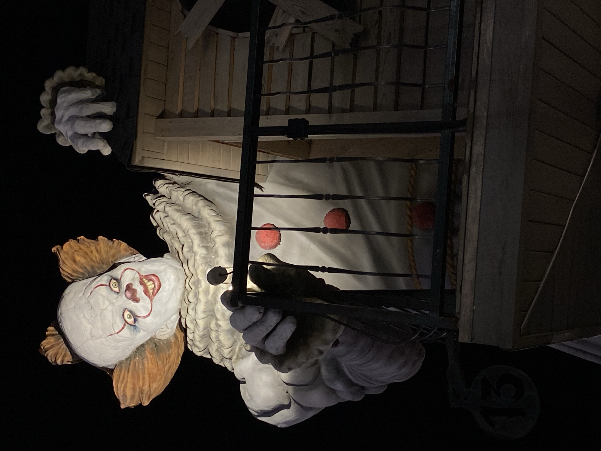 The image shows a large, menacing clown statue, resembling Pennywise from Stephen King's "It," positioned on the exterior of a building at night. The statue is illuminated, highlighting its white, clown-like face with red markings around the eyes and mouth, and a wide, unsettling smile. Its large, puffy costume is visible, as well as its hands. One hand is gripping the railing of a balcony, and another hand appears to be emerging from the roof of the structure. The building itself appears to be constructed of wood and has a dark, sloped roof. A circular sign with the number "13" is visible on the left side of the statue. The setting is outdoors, with a dark, black background indicating nighttime. The statue is positioned at an angle, giving a sense of its imposing size and a slightly disturbing perspective. There are no people visible in the image. The statue is part of an exhibit or attraction in Gelendzhik, Russia.