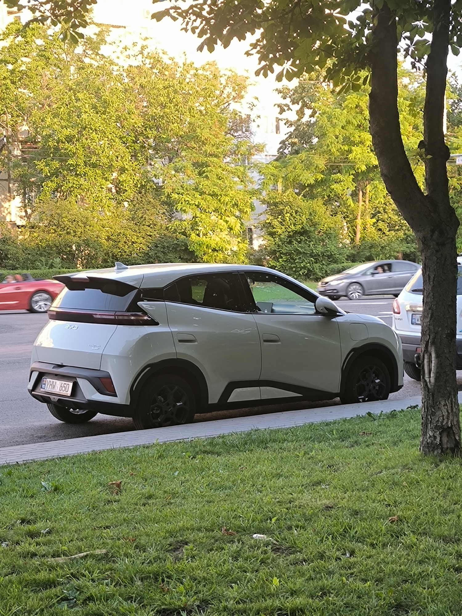 The image captures a white BYD Yuan UP electric vehicle parked on the side of a street in Chișinău, Moldova. The car is positioned facing right, with its rear visible. The license plate displays "YHM 650". The scene is set in a tree-lined urban environment with lush green grass and foliage. In the background, other vehicles are present, including a red sports car in motion and a grey sedan, indicating a street with active traffic. The lighting suggests it is late afternoon or early evening, with sunlight filtering through the leaves of the trees. The overall impression is a snapshot of everyday urban life in Chișinău.