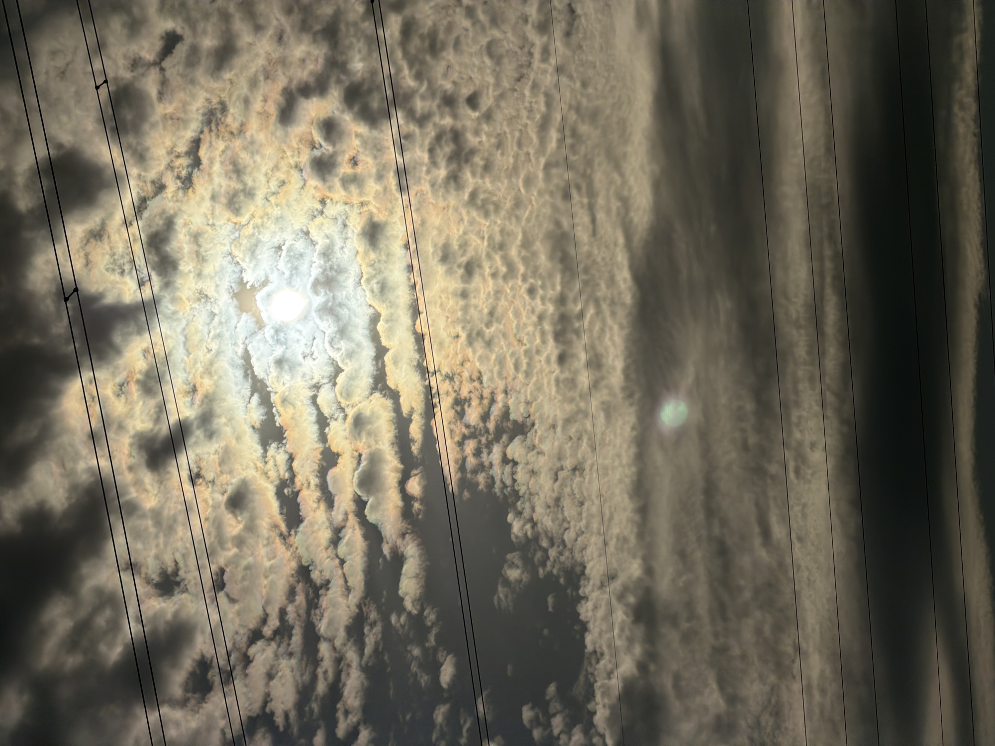 The image shows a cloudy sky with the sun shining through, creating a bright halo effect. Several power lines are visible, stretching horizontally across the frame. The clouds are varied, with some appearing dark and heavy, while others are lighter and illuminated by the sun. There are no people or specific landmarks visible that would indicate a location in Minsk, Belarus. The scene is purely atmospheric.