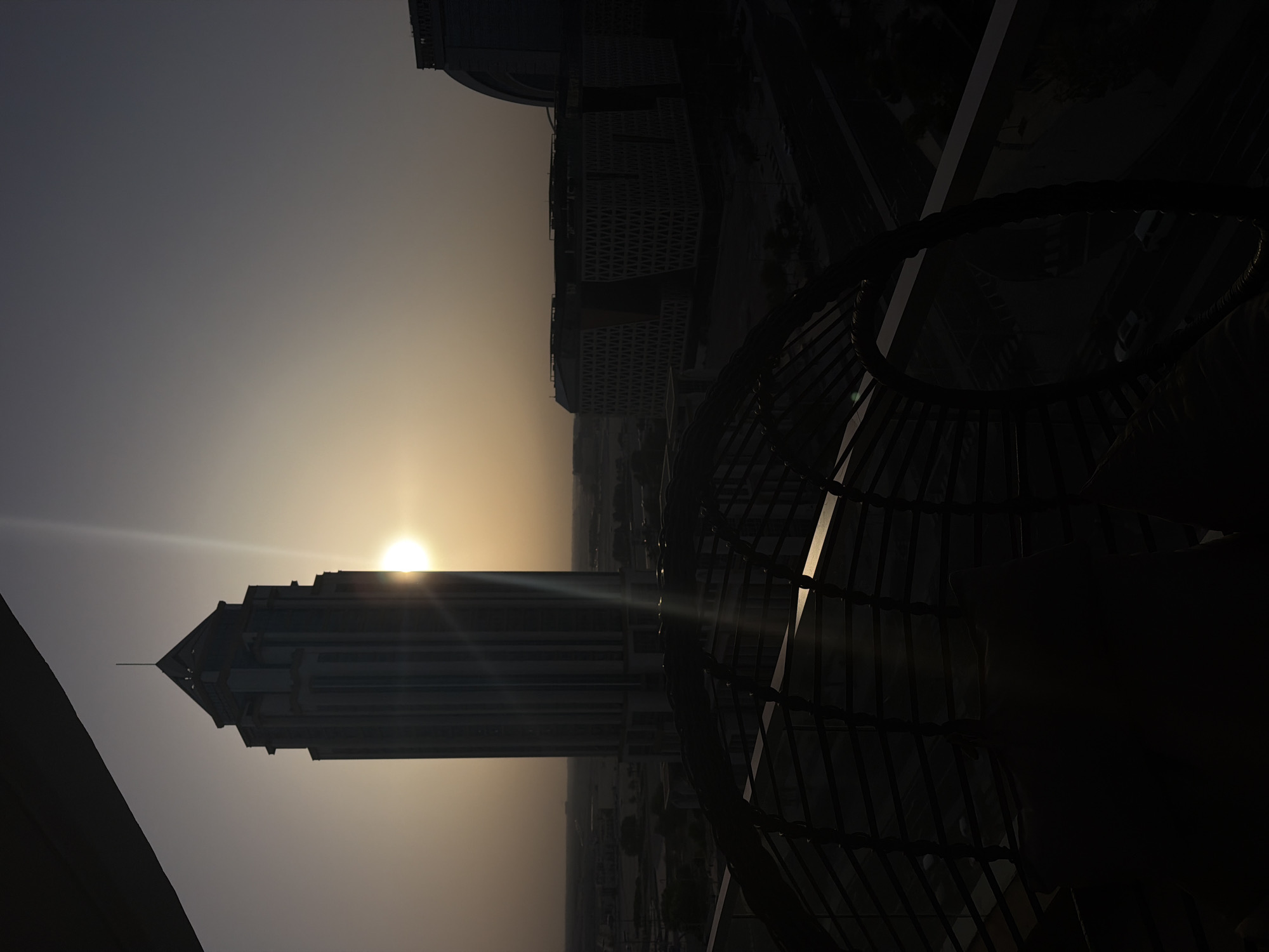 This media file captures a striking sunset over a modern cityscape, likely Lusail, Qatar. The sun, a brilliant orb, is partially obscured by a tall, geometric skyscraper, casting a lens flare that streaks across the frame. The skyscraper, with its stepped facade and pointed apex, is silhouetted against the warm, gradient sky, which transitions from a pale yellow near the horizon to a dusky gray above.
In the foreground, a dark, ornate metal structure, possibly a decorative screen or railing, frames the scene on the right. Its intricate pattern creates a sense of depth and adds a contrasting element to the sleek lines of the architecture. The overall impression is one of urban grandeur meeting the dramatic beauty of a sunset.
The time of day is clearly evening, with the low-angled sunlight indicating sunset. The atmosphere appears clear, allowing for the sun's rays to be sharply defined. The image evokes a sense of tranquility and awe, highlighting the striking architecture and the natural spectacle of the sunset. No text is visible in the media file.