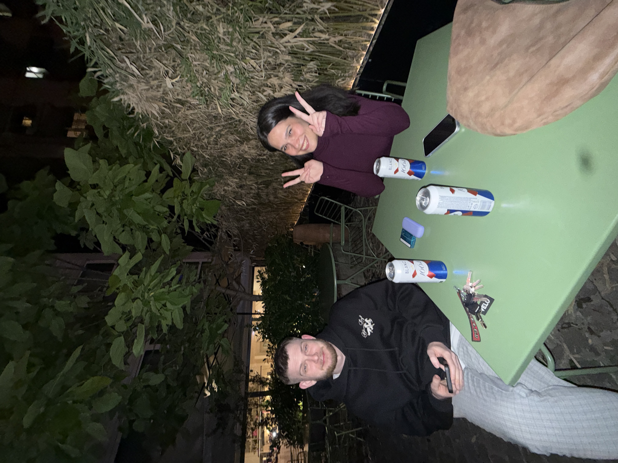 Two people are seated at a square, green metal table outdoors at night. A woman with long dark hair is making a peace sign. She is wearing a dark purple long-sleeved shirt. A man is wearing a black hooded sweatshirt and gray pants. Three cans of beer labeled "Blanc 1664" are on the table, along with a cell phone, car keys, a small pack of gum, and what appears to be a photograph or a small postcard. The setting looks like an outdoor bar or restaurant in an urban area, with foliage in the background and buildings visible in the distance. Based on the outdoor metal furniture, type of foliage, beer brand, and visible architecture, it is likely in Belgrade, Serbia.