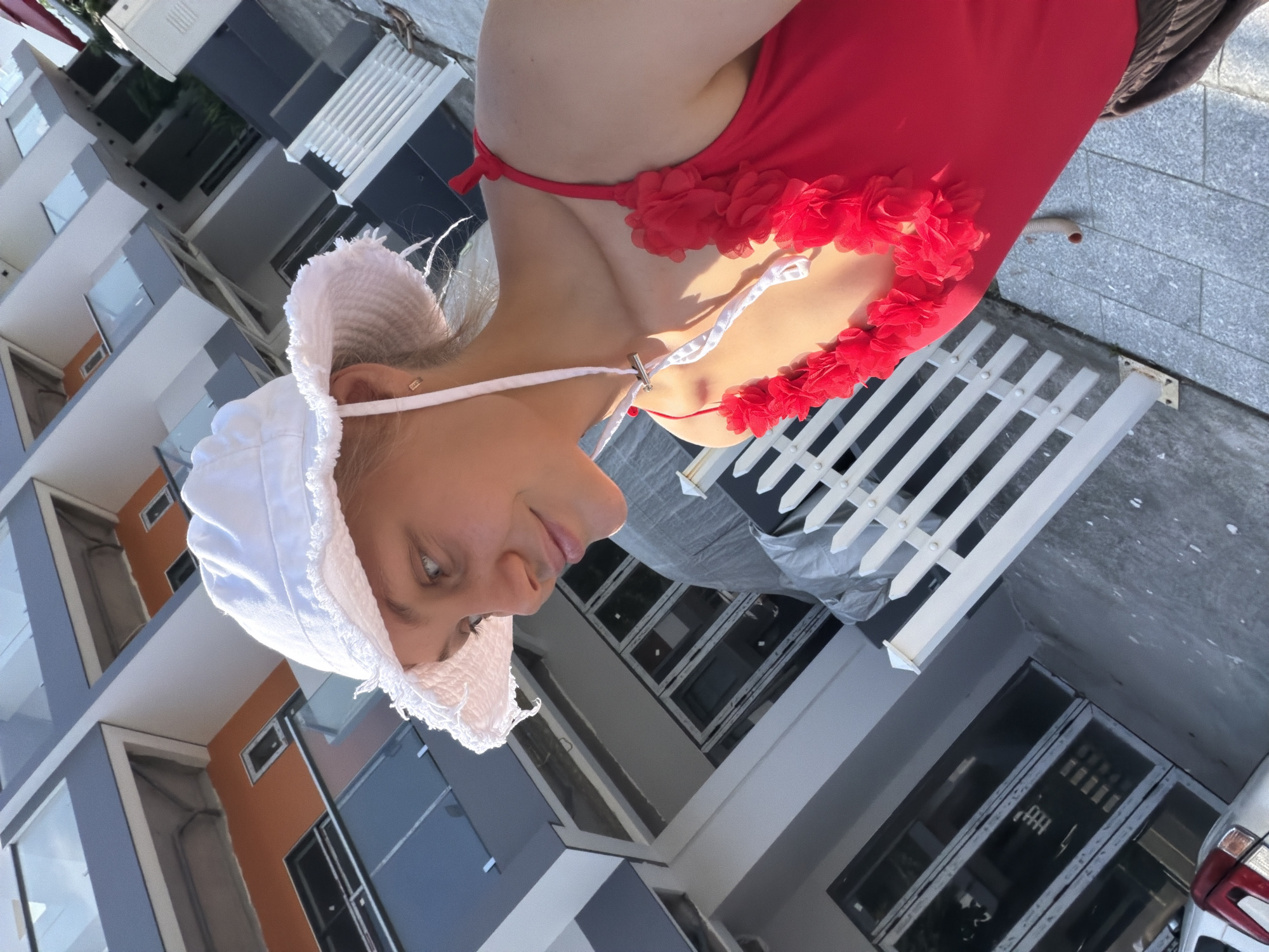 A young woman, wearing a red swimsuit with floral embellishments and a white sun hat, stands outdoors in what appears to be a construction or renovation zone. The background features a modern building with gray, orange, and blue facades, and large glass windows. A white picket fence is visible in the foreground, and to the left, part of a silver vehicle is present. The ground is paved with grey tiles. The woman is looking upwards, with a slight smile. The setting suggests a location within Nam Nha Trang Ward, Vietnam, possibly a residential or commercial development area.