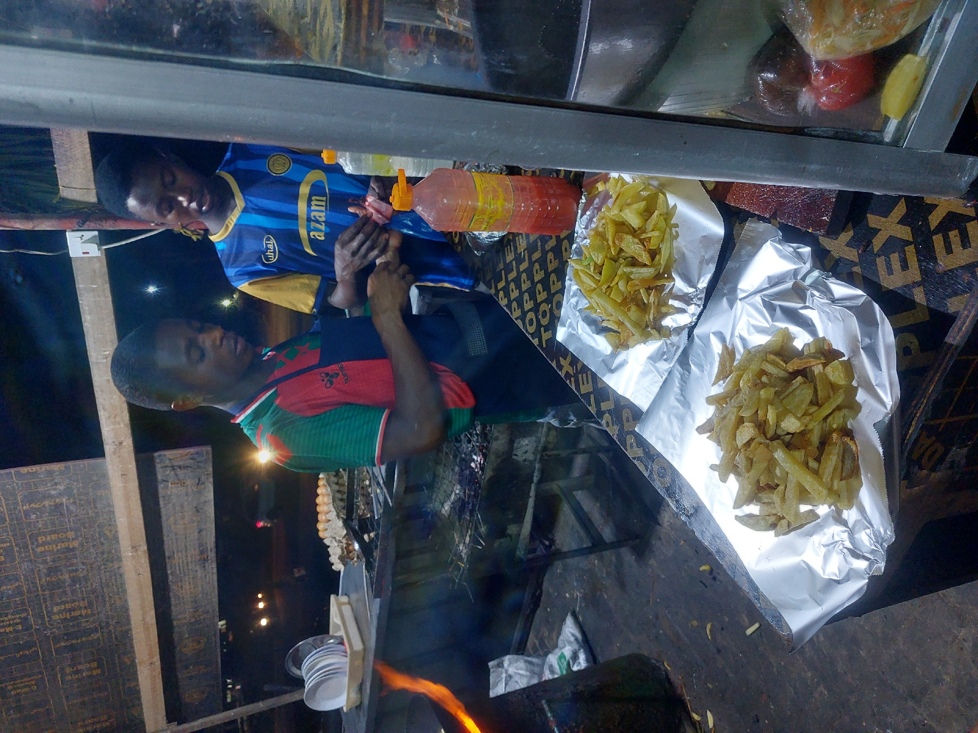 The image shows a roadside food stall at night, likely in Dar es-Salaam, Tanzania. Two young men are present. One wears a blue and yellow Azam FC jersey, while the other wears a green, red, and black jersey. They are standing behind a food preparation surface. On the surface are two piles of French fries served on aluminum foil, and a large bottle of red sauce. Behind the preparation surface is a charcoal grill with flames visible. A stack of white plates is attached to the side of the grill. In the background, the city lights are visible. A string of eggs are hanging down. The stall appears to be constructed from plywood, with the word "PLEX" visible.