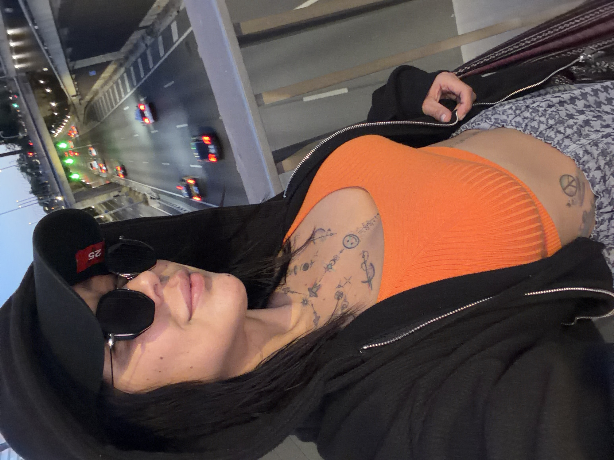 A woman wearing a black hoodie, sunglasses, and an orange top is pictured in a high-angle, medium shot. She has tattoos on her left arm and side. To the left of the woman, there is a bridge and road with cars on it. The location is in L'Hospitalet De Llobregat, Spain.