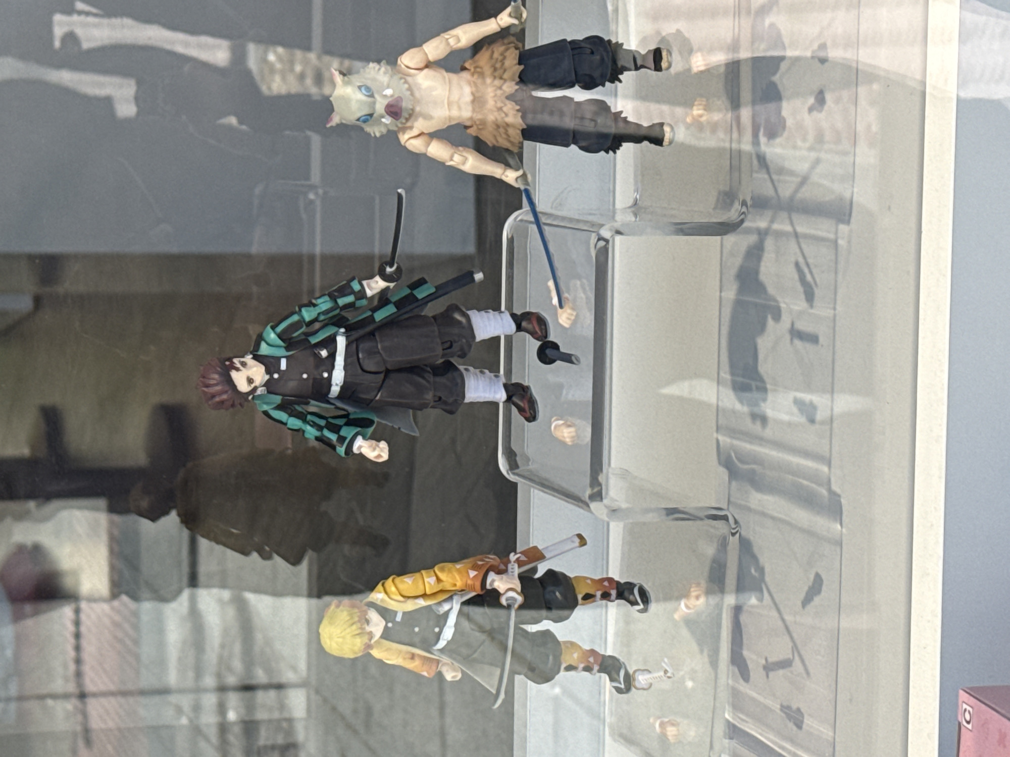 The image shows three anime figurines on clear shelves, likely inside a display case. The top figurine appears to be Inosuke Hashibira from Demon Slayer, characterized by his boar mask. The middle figurine is Tanjiro Kamado, also from Demon Slayer, recognizable by his green and black checkered haori and sword. The bottom figurine is Zenitsu Agatsuma, another Demon Slayer character, featuring his yellow and orange patterned haori and sword. The background reflects a cityscape, possibly taken in Madrid, Spain, given the architectural style and light. Reflections are visible on the glass display case, obscuring some details of the surrounding environment.