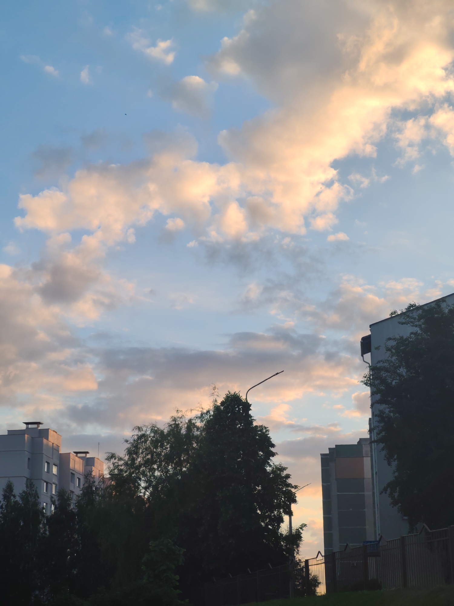 The image shows a view of several buildings in an urban environment. The sky is filled with clouds, some of which are illuminated with a warm, golden light. Buildings are partially obscured by trees. A street lamp is visible in the middle ground. A fence is at the bottom. The location appears to be in the city of Unknown, Unknown.
