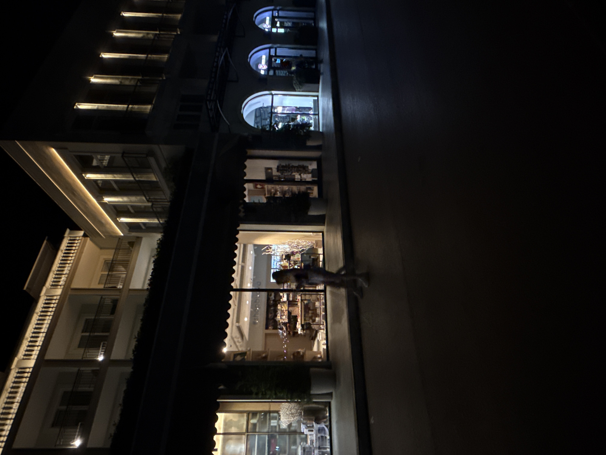 The image captures a street scene at night in Budva, Montenegro, likely along a pedestrian walkway or promenade adjacent to shops and buildings. The lighting is dim, with the main illumination coming from the illuminated windows of businesses and decorative lights on the buildings.
In the foreground, a paved street or walkway is visible, reflecting some of the ambient light. A person, appearing as a blurred silhouette, is walking on this walkway. Their attire is indistinct due to the low light conditions.
The middle ground features the facades of buildings, which appear to be a mix of commercial establishments and residential or hotel units above. Several storefronts are visible, showcasing interiors with displays of merchandise. One prominent storefront has large glass windows, revealing shelves stocked with items. Another section of the building features arched doorways leading into businesses, with some displaying colorful lighting. The architectural style appears modern with classical influences, evident in the balconies and decorative elements on the upper floors.
The background consists of the upper levels of the buildings, which are illuminated by soft, warm lighting, highlighting architectural details such as railings and window frames. The overall impression is one of a well-lit commercial or tourist area at night.