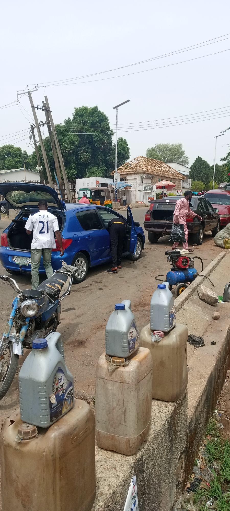The image depicts a roadside scene in the city of Jos, Nigeria. Several cars are parked alongside the road. A blue hatchback has its rear door open, and a person is working on it. A man wearing a white jersey with the number 21 stands nearby. Another person is working on the driver's side of the car. To the right, a dark-colored car is parked, with a man in a pink outfit appearing to interact with it. An air compressor sits on the ground near this car. In the foreground, several large, dirty containers hold Mobil Super 1000 motor oil. A motorcycle is also parked in front of these containers. In the background, there is a building undergoing construction with only the roof frame completed. A tricycle taxi and a small shop labeled "POS" can be seen. Utility poles and power lines are visible in the background. The overall environment appears urban, slightly cluttered, and possibly a car repair or maintenance area.
