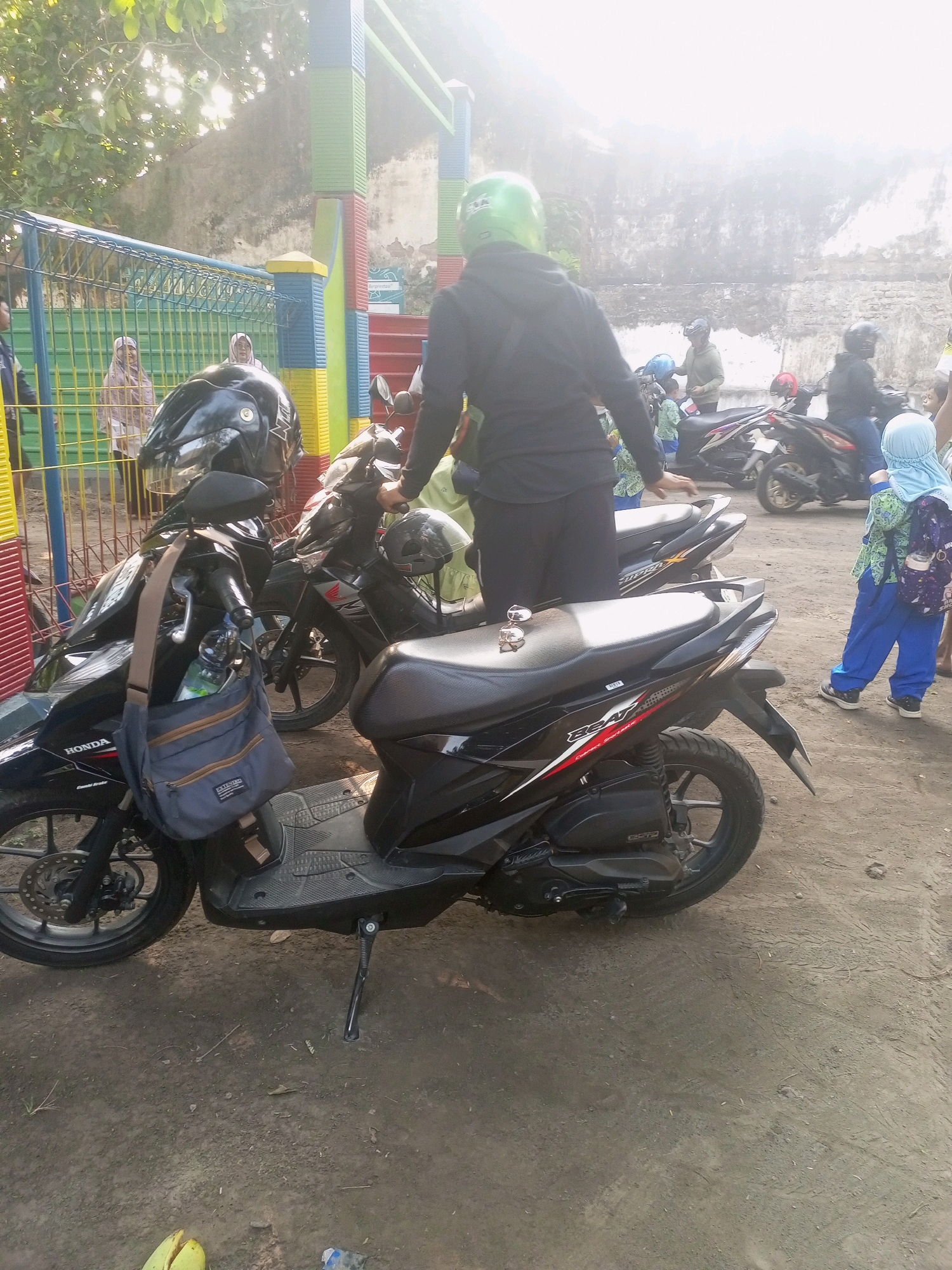In Gurah, Indonesia, a street scene unfolds with multiple motorcycles parked outdoors on a dusty ground. In the foreground, a black Honda scooter is prominent, equipped with a side bag and a water bottle. Behind it, another black motorcycle is visible. Several people are present: a person wearing a black jacket and a green helmet stands near the scooter, facing away from the camera. To the right, a child in blue pants and a hijab with a backpack walks away from the viewer. Other individuals, including those wearing hijabs, are partially visible in the background, some near other motorcycles. The environment includes a fence made of metal bars and colorful vertical posts in shades of blue, green, yellow, and red. A weathered wall forms the backdrop, and trees with sunlight filtering through their leaves are visible. The lighting suggests it is daytime, possibly late afternoon due to the quality of light.